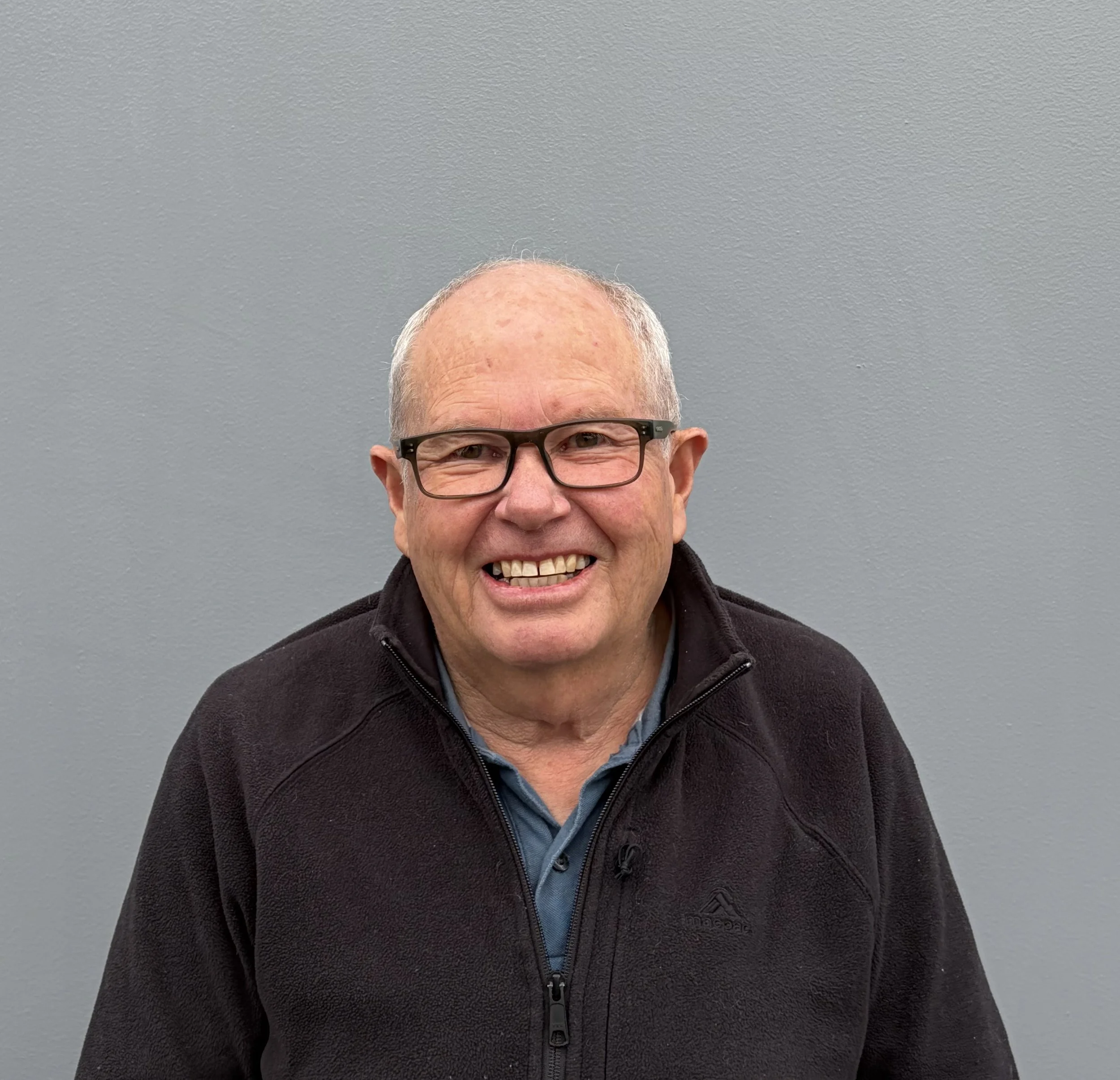 Close-up of a smiling elderly man with gray hair, glasses, wearing a black zip-up jacket over a blue shirt, standing in front of a gray wall.