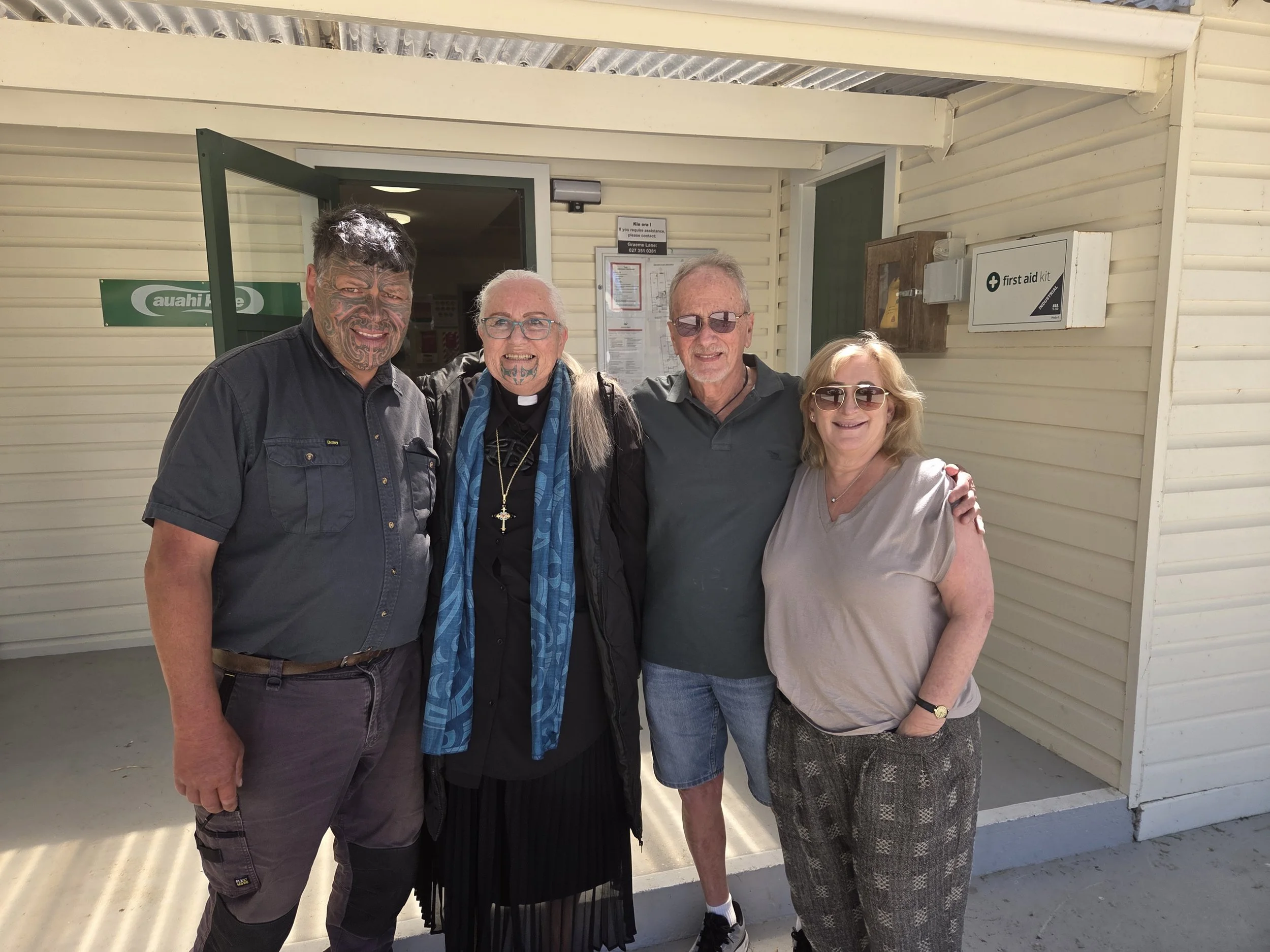 Four people standing outside a building with beige siding, smiling at the camera. The group includes two men with short gray or dark hair, a woman with blonde hair and sunglasses, and a religious woman with a cross necklace and glasses. The building has signs, one indicating an aahliite pharmacy and another for a first aid kit.