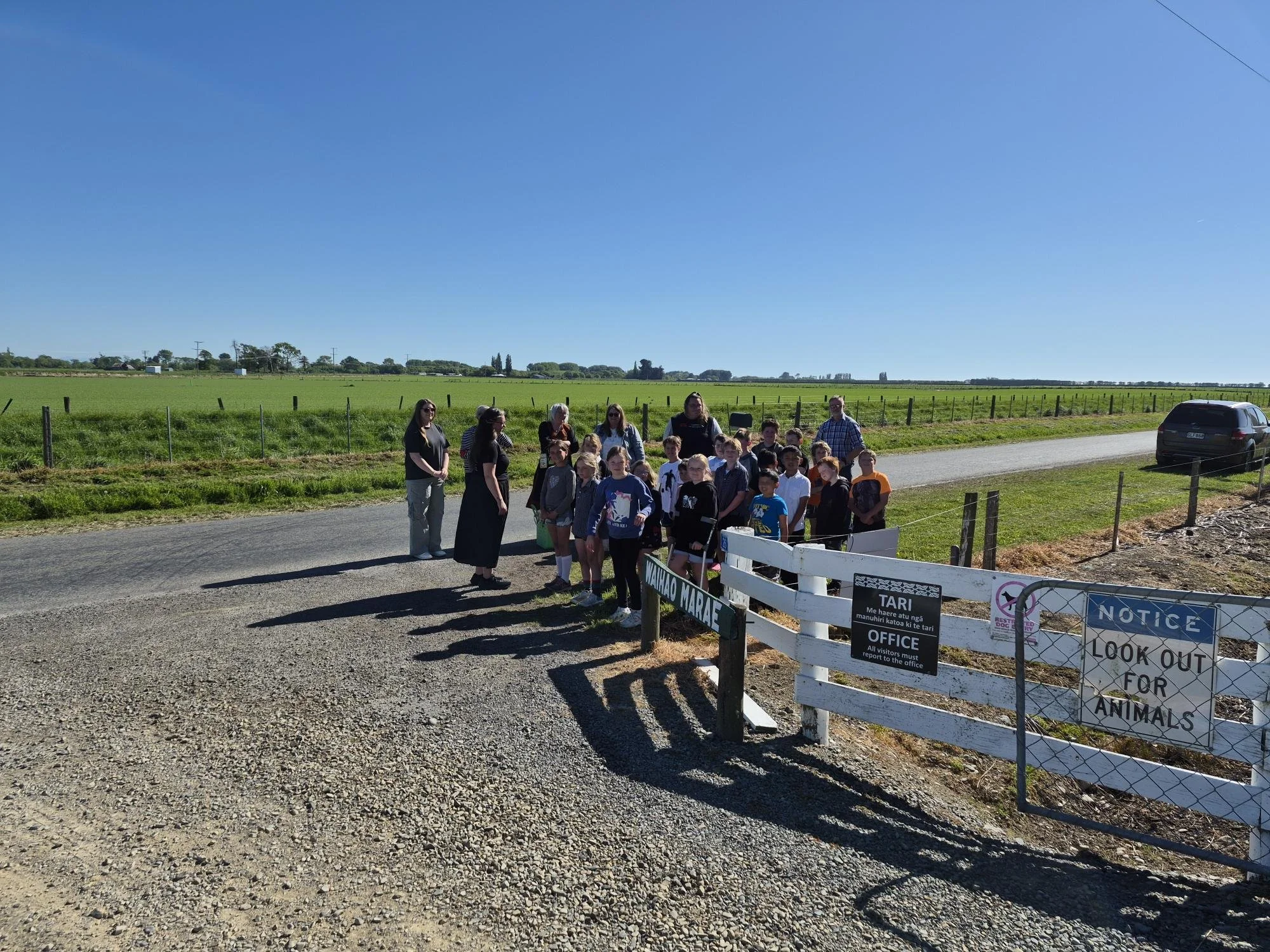 A group of children and adults standing outside in front of a white fence, near a sign that says 'NOTICE' and 'LOOK OUT FOR ANIMALS'. There is a gravel road in the foreground and open green fields under a clear blue sky in the background.