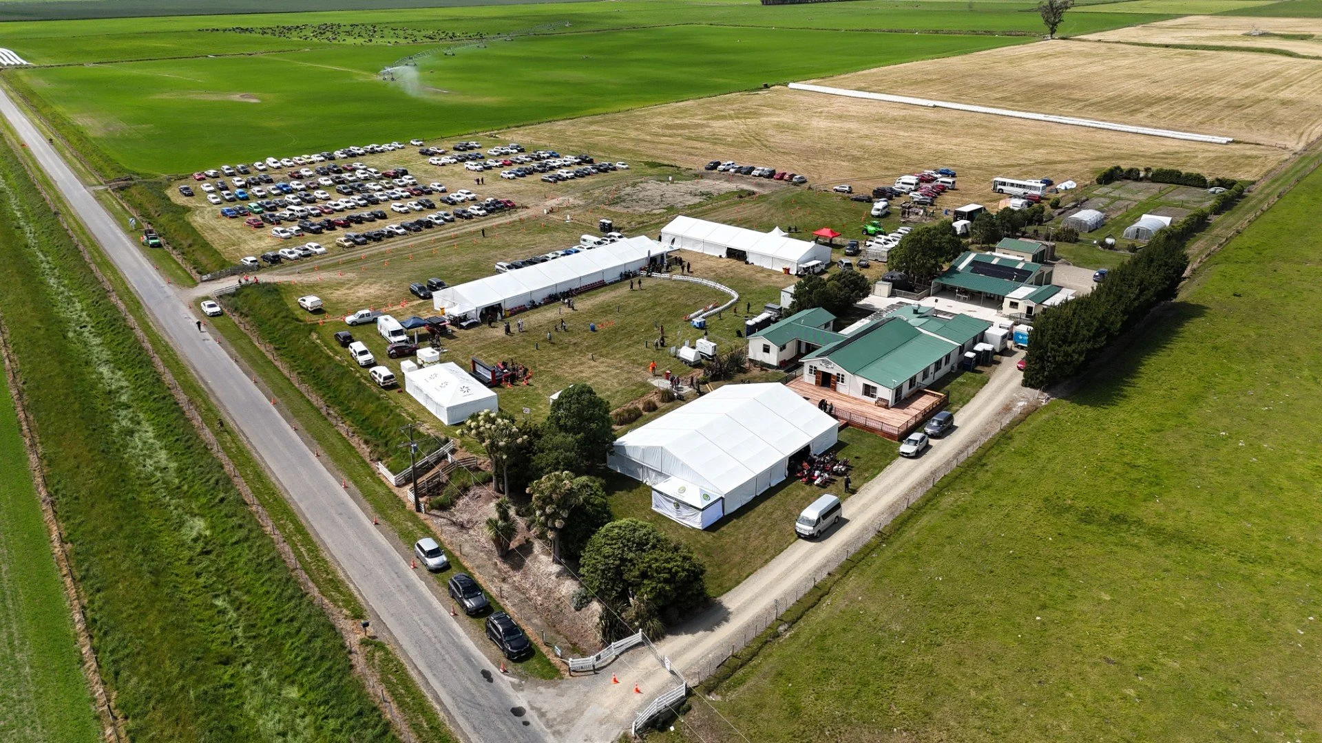 An aerial view of a farm-like venue with multiple white tents, a large parking lot filled with cars, and a long driveway. The area has green fields around it, with some farm structures and greenhouses visible.