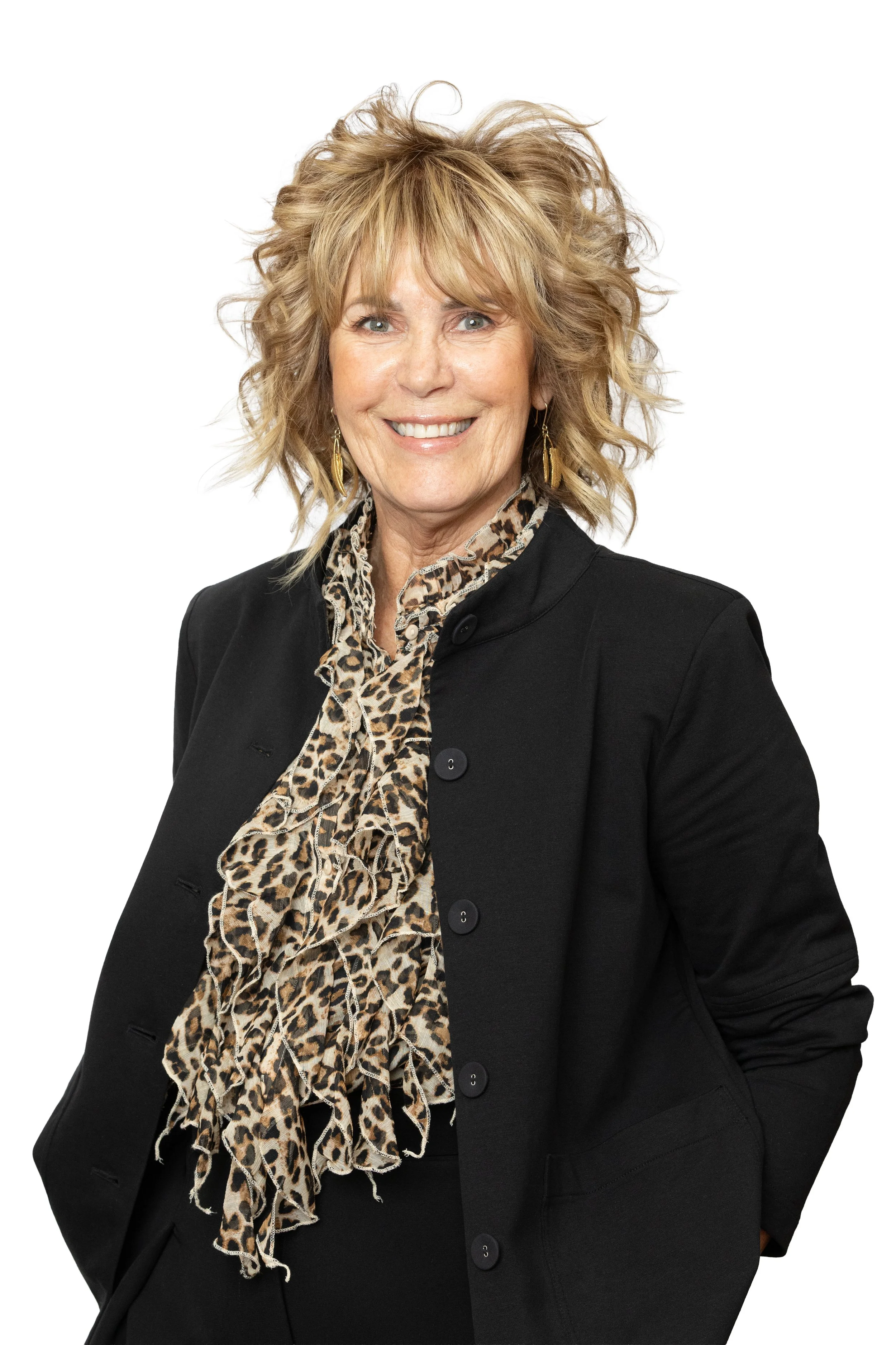 Portrait of a smiling middle-aged woman with blonde, curly hair, wearing a black blazer and a leopard print blouse with ruffles, gold earrings, against a white background.