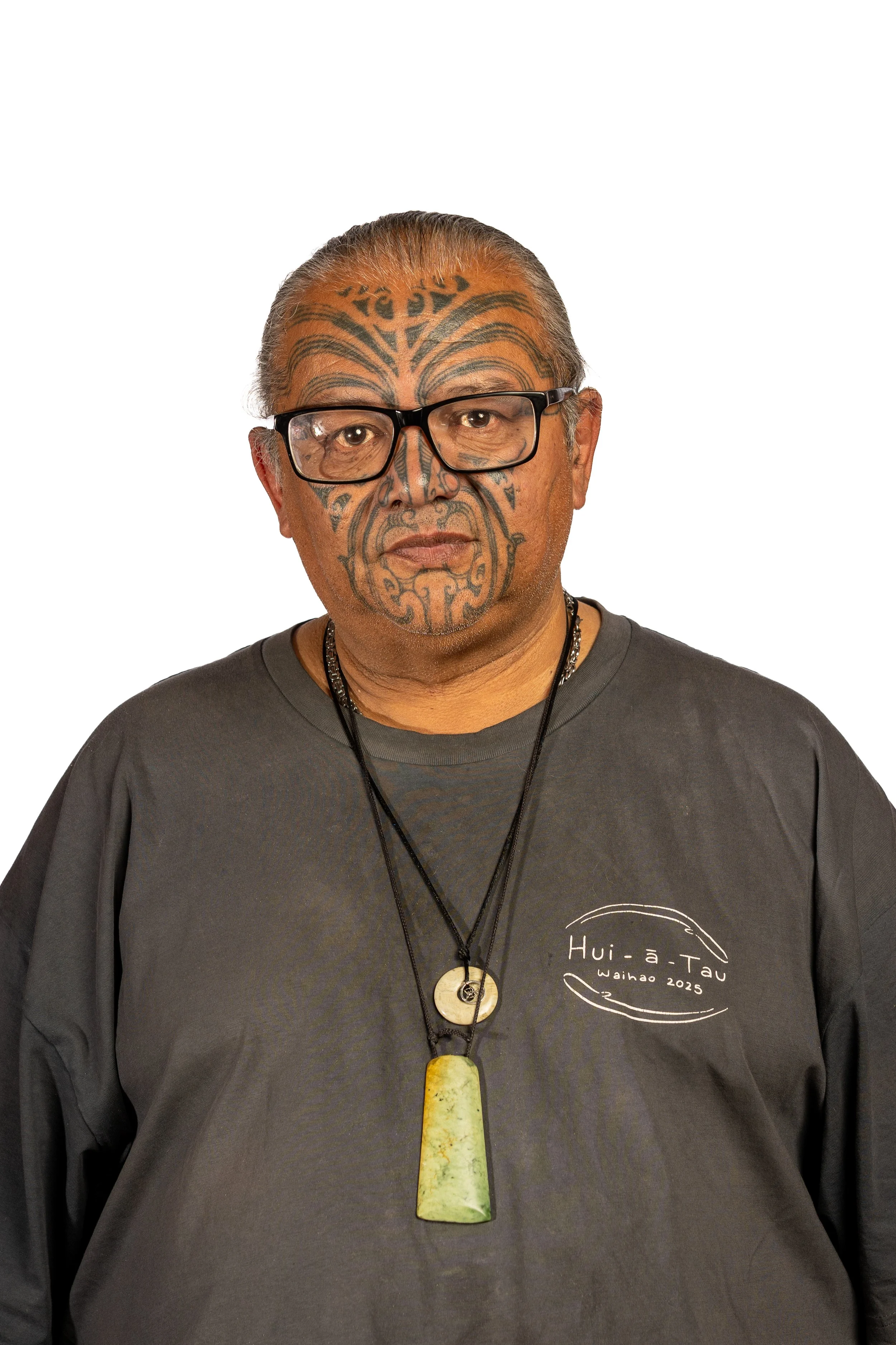A man with glasses and gray hair, wearing a gray shirt with white text and graphics, and a necklace with a large greenish pendant. He has traditional Polynesian tattoos on his face and head.