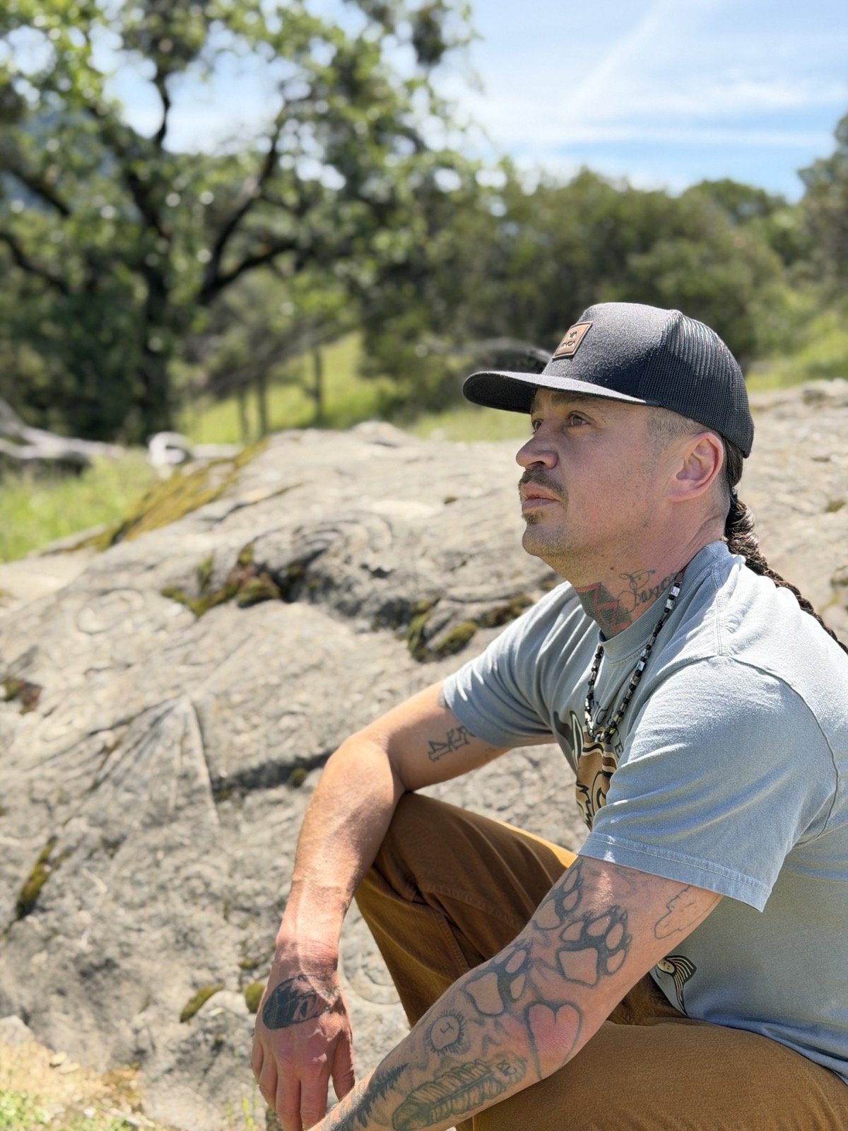 A man sitting on a rock outdoors with trees and blue sky in the background, wearing a black cap, gray T-shirt, and brown pants, with tattoos on his arms and neck.