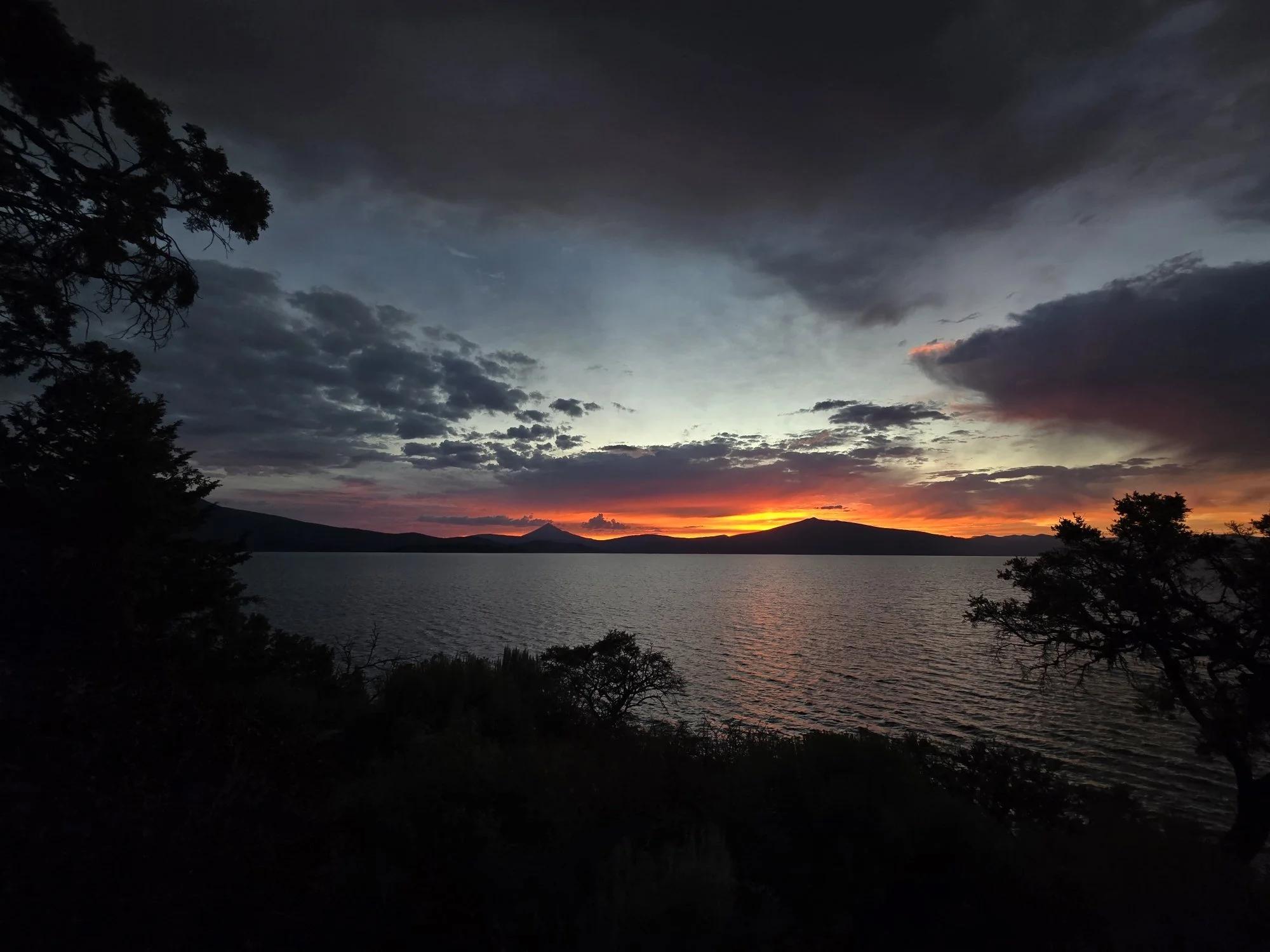 Sunset over a body of water with silhouetted trees and distant mountains, some dark clouds in the sky.