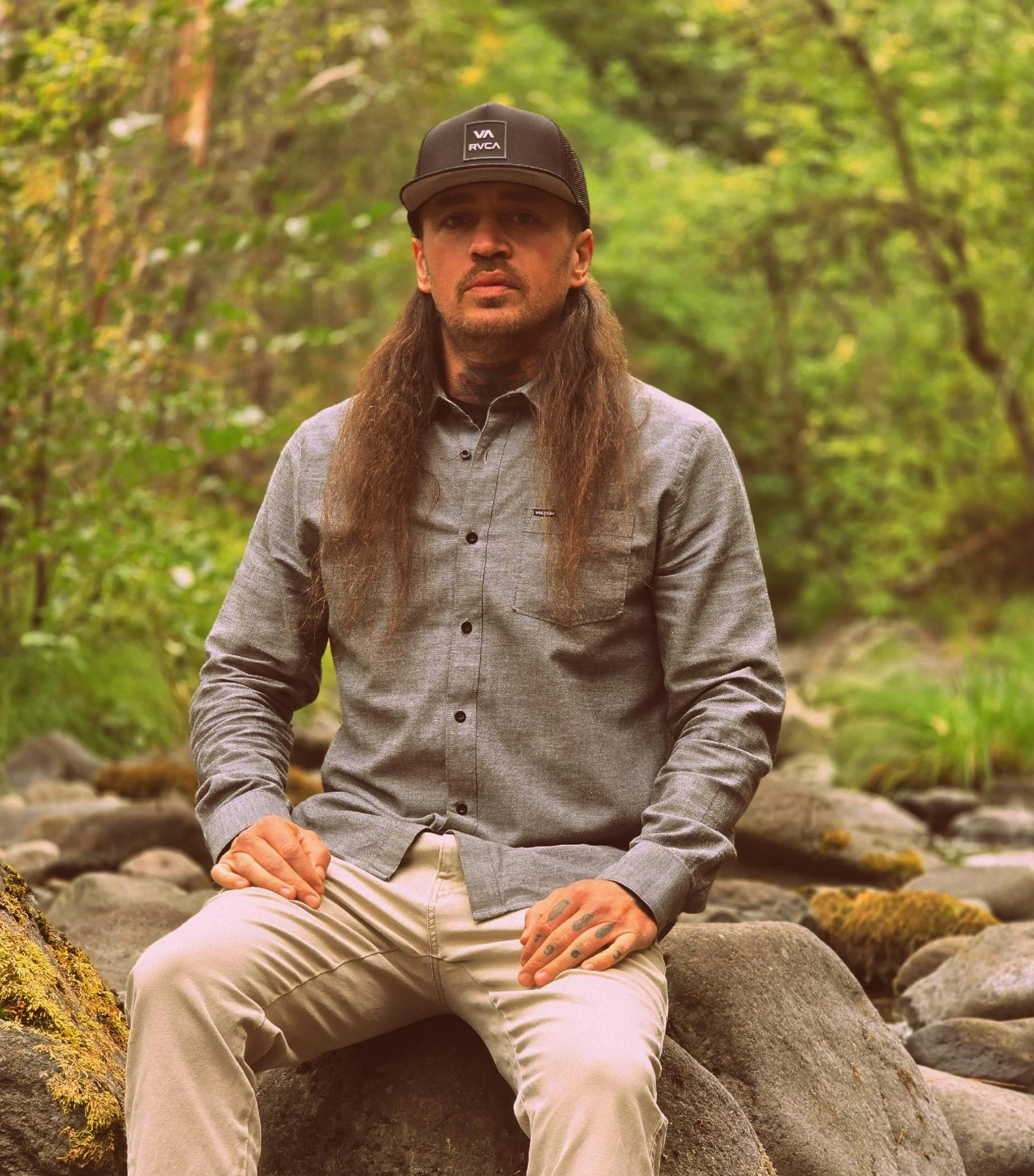A man with long hair, beard, and tattoos on his fingers is sitting on a large rock in a wooded area with green foliage, wearing a gray button-up shirt, beige pants, and a black cap.