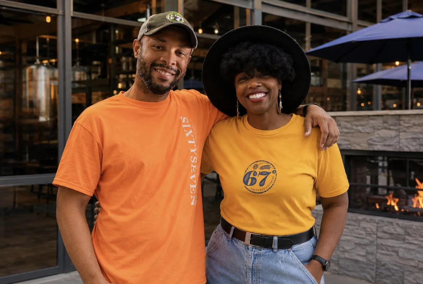 Two smiling people, a man and a woman, stand together with their arms around each other outdoors in front of a fire pit and wooden structure, dressed casually in yellow and orange T-shirts.