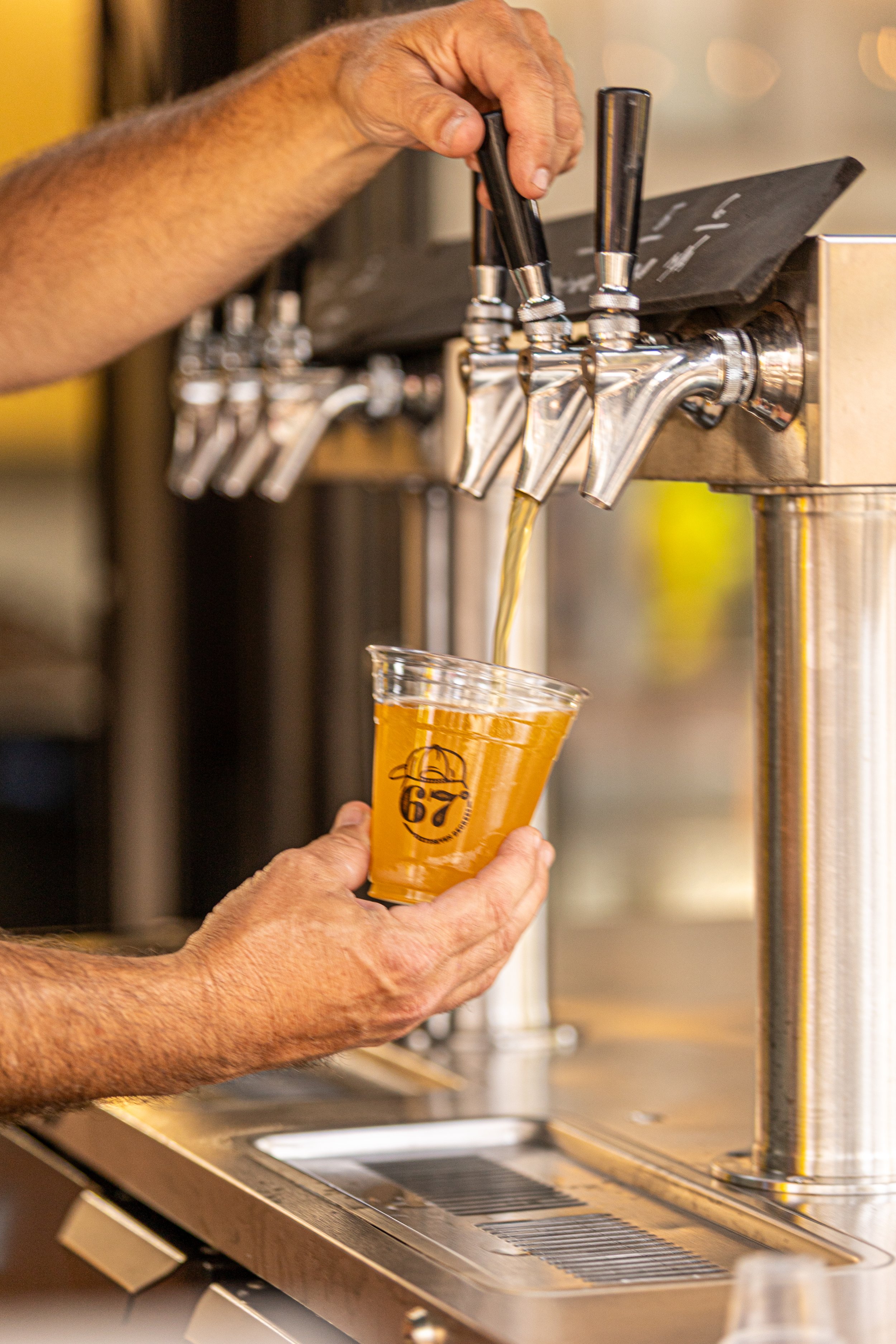 A person with tan skin and visible arm hair is pouring beer from a tap into a clear plastic cup at a bar or brewery. The beer is golden in color and the person's hand is holding the cup with a logo on it. The background shows the beer taps and a metallic serving station.