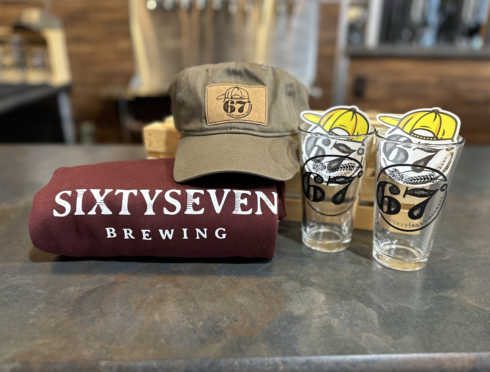 A maroon cloth with white text reading 'SixtySeven Brewing' on a dark surface. A khaki baseball cap with a '67' patch is placed on top of the cloth. Two clear glasses with the 'SixtySeven Brewing' logo, each with a paper tag featuring a cartoon beer with a yellow hat, are beside the cloth.