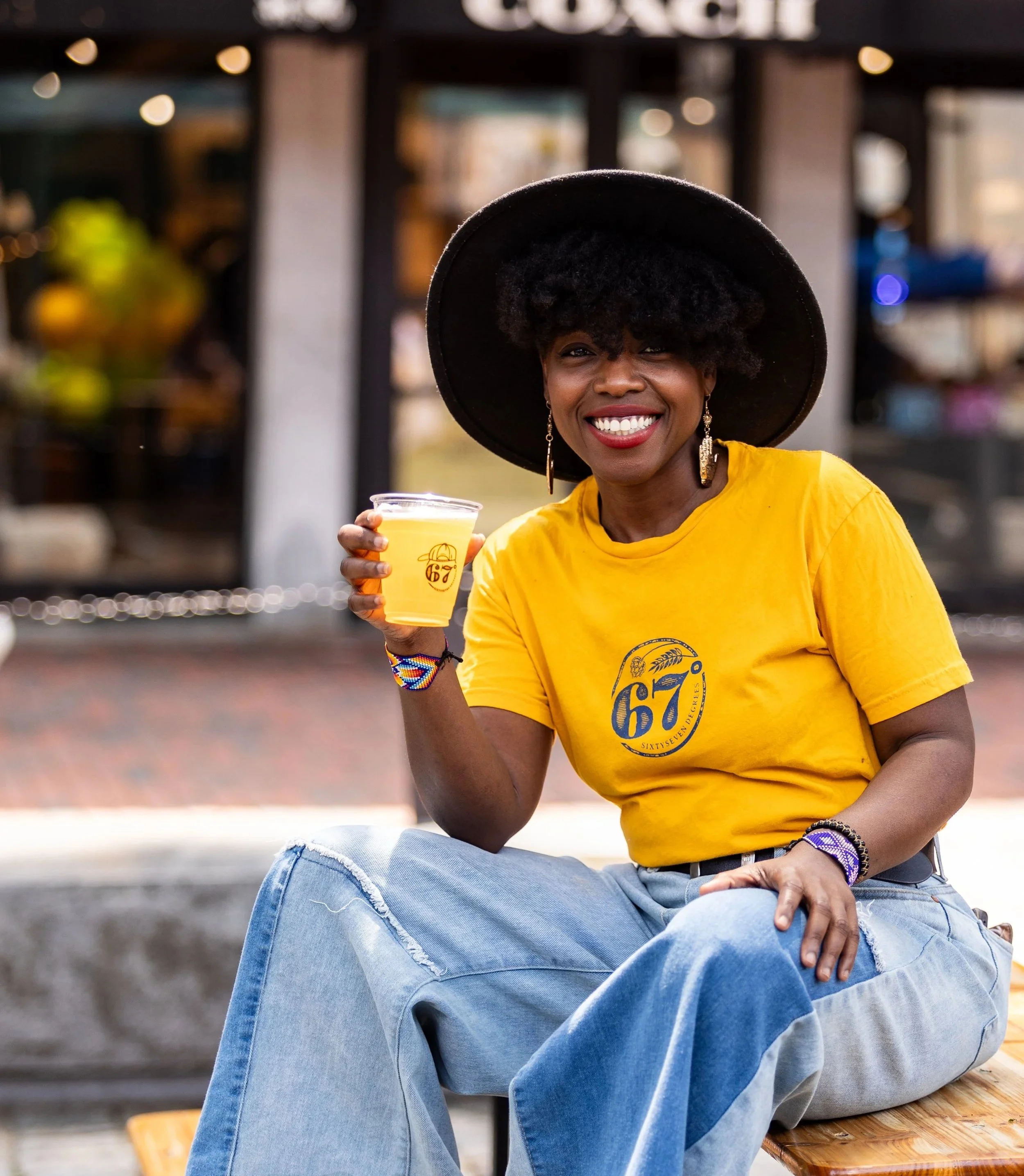 A woman with dark skin and curly hair is smiling while sitting outdoors on a wooden bench, holding a yellow drink. She is wearing a large black hat, a yellow T-shirt, and ripped blue jeans. The background is blurred with lights and other urban elements.