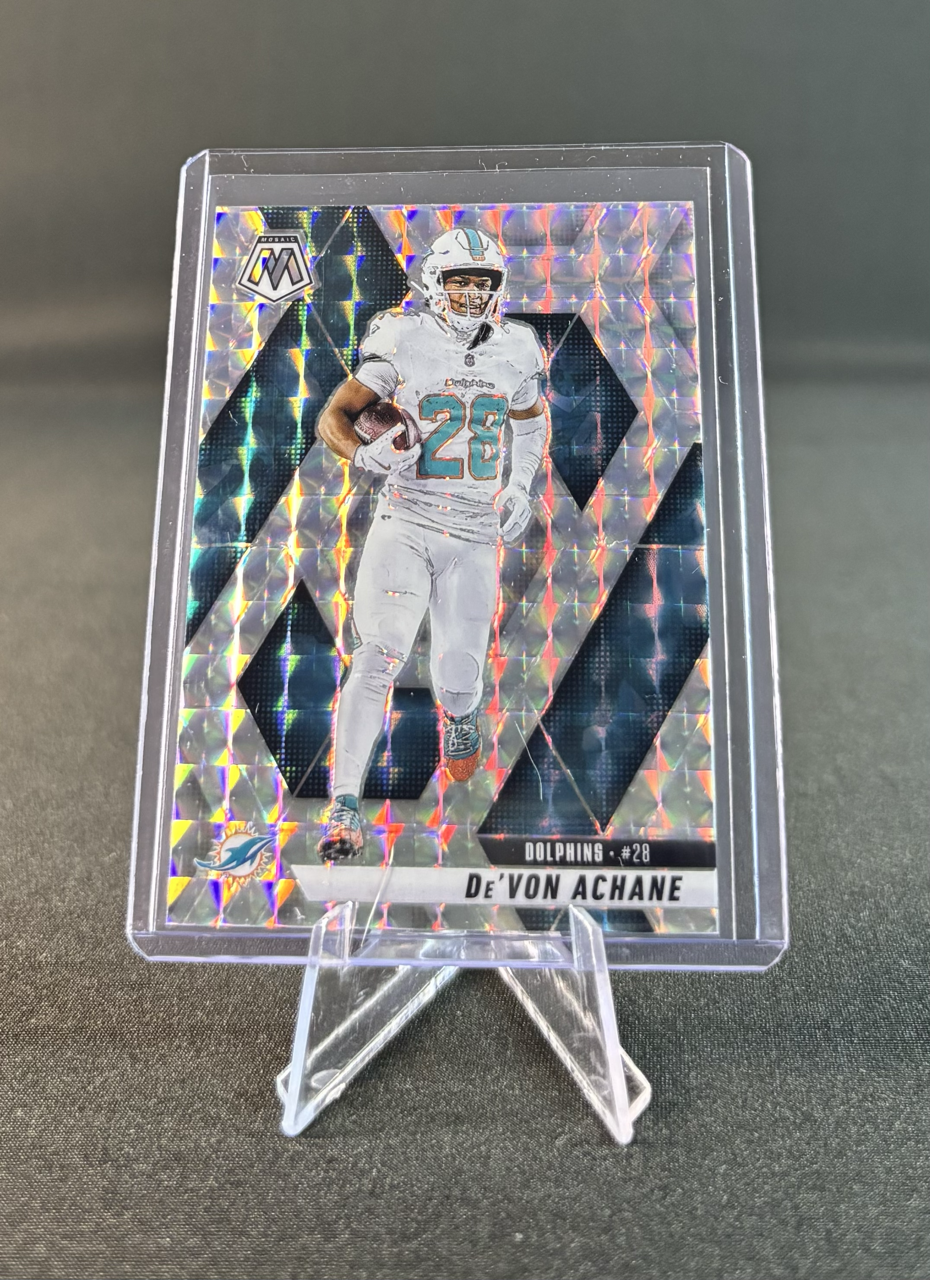 Holographic trading card of football player De'Von Achane in Miami Dolphins uniform, holding a football, standing on a clear acrylic display stand.