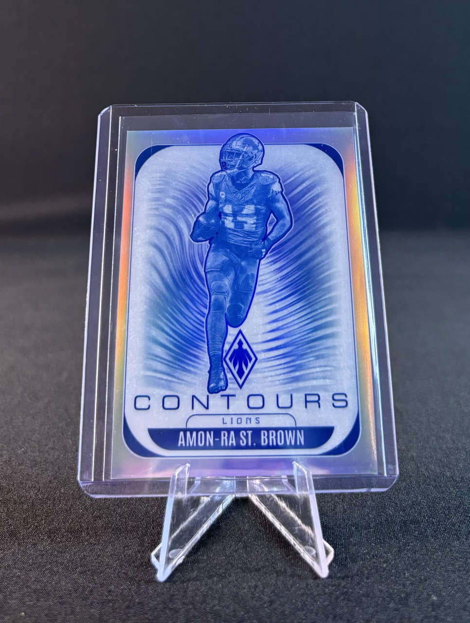 A holographic football trading card featuring a player in a blue uniform running forward, with text indicating the player is Amon-Ra St. Brown from the Lions team, branded 'Contours'.