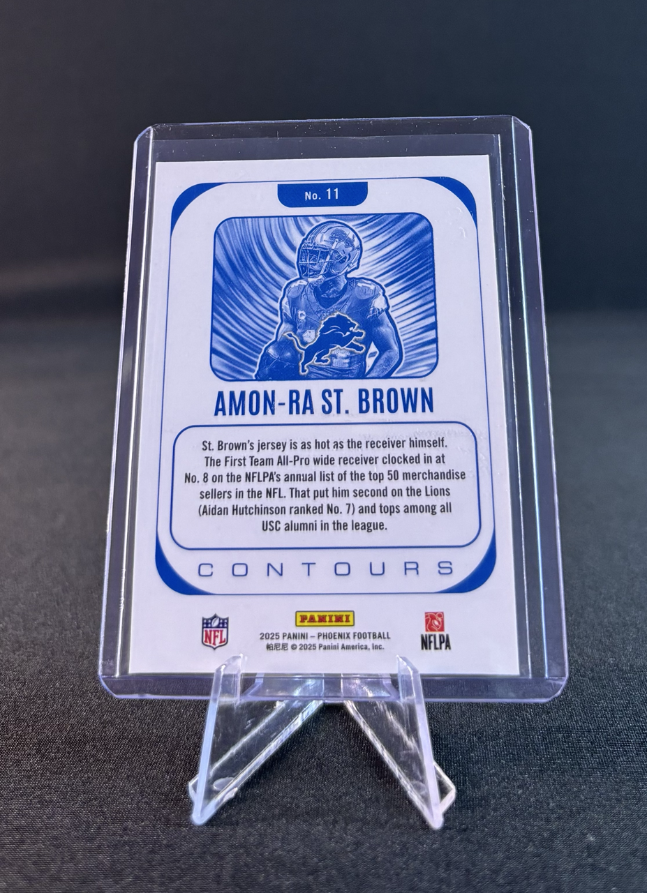 A football card featuring Amon-Ra St. Brown with a blue illustration of him in his Detroit Lions uniform carrying a football, surrounded by a blue background with radiating lines. The card includes text about his jersey and ranking, and logos of NFL, Panini, and NFLPA.