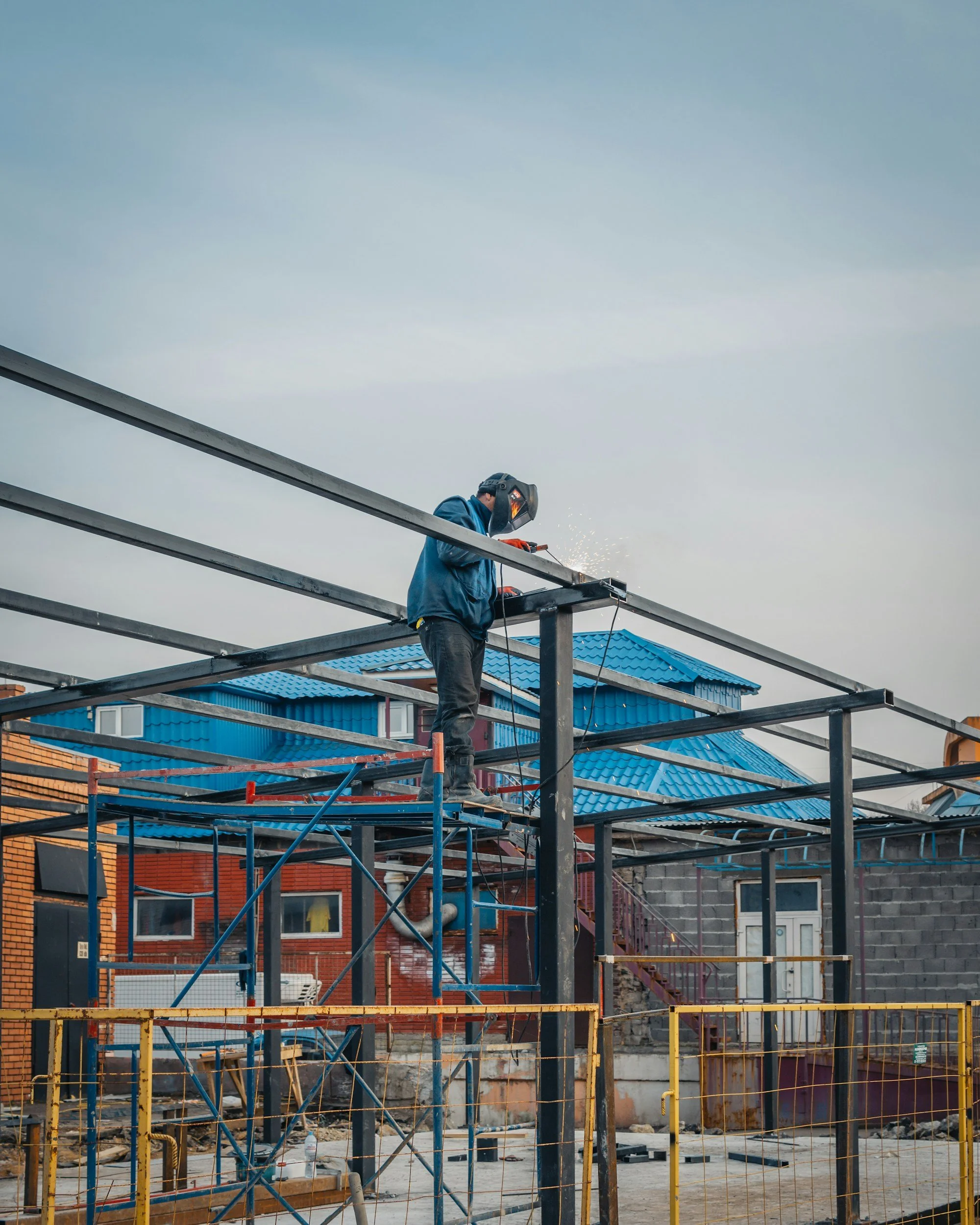 A construction worker welding steel beams on a building site with a blue house and other buildings in the background.