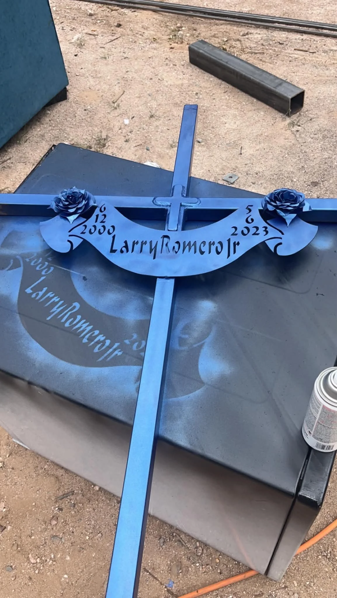 Blue metal cross with decorative blue roses and the text 'Larry Romero 12 15 2000 5 6 2023' in the center. The cross is placed on a metallic surface outdoors.