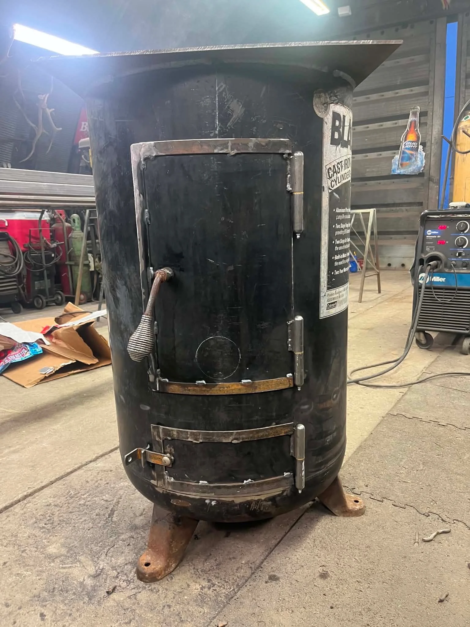 A large black industrial oven or smoker with rusted legs is located in a workshop or garage. The back of the oven features a door and exhaust pipe, with a piece of tape or sticker on the side. The surrounding area includes tools, a welding machine, cardboard, and a workbench with equipment.