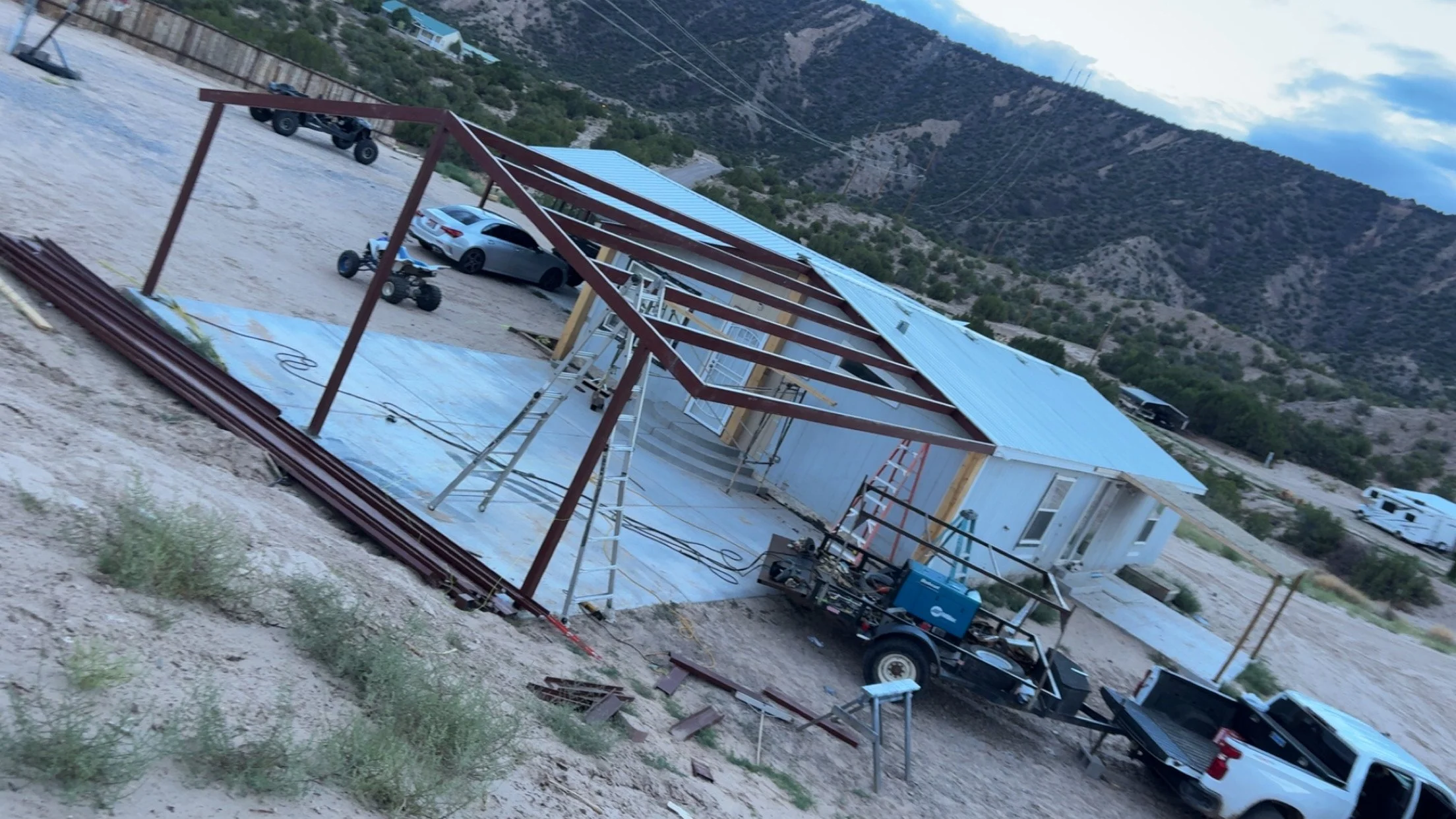 Construction site with a building under construction, a red steel frame, and a silver car parked nearby, with mountains in the background.