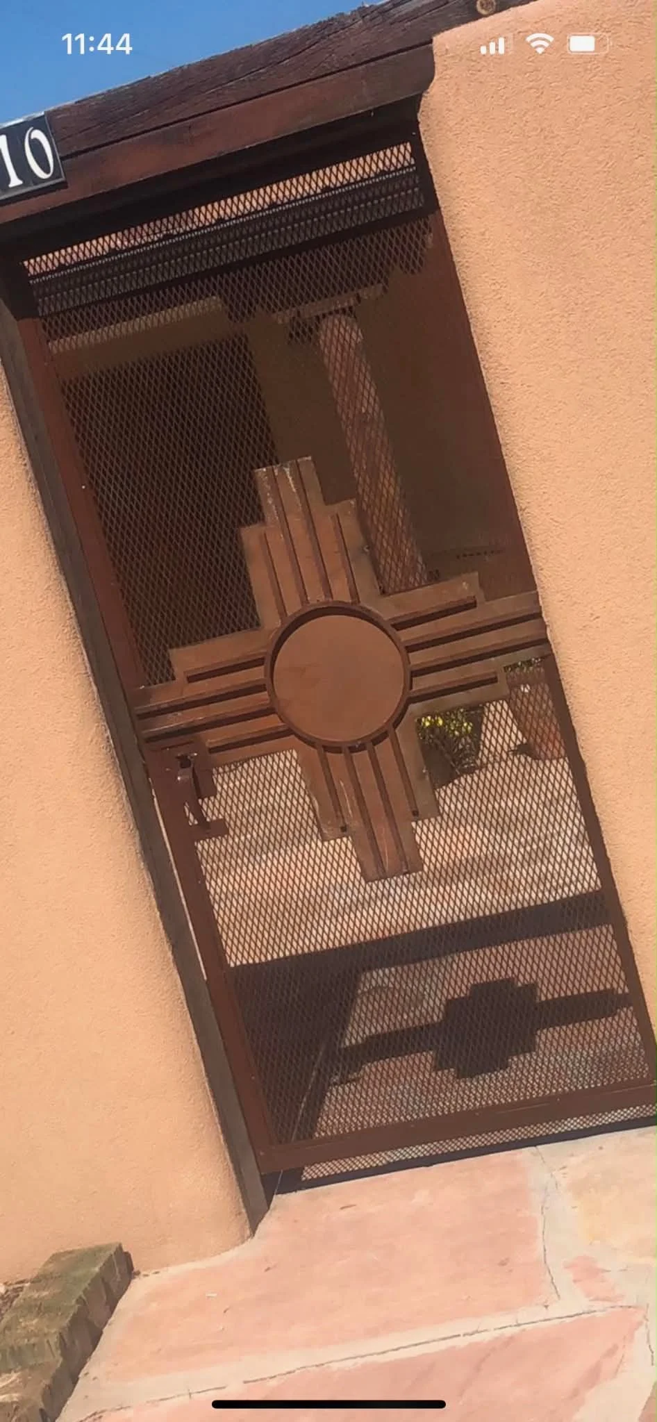 A metal security gate with a decorative cross design and wooden doors behind it, set against a beige stucco wall with a stone ledge at the bottom.