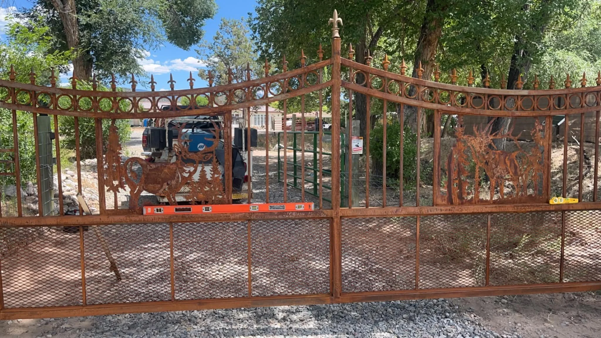 Rusty iron gate with decorative points and tree-shaped designs, partially open to a yard with trees and a gravel ground. A truck with equipment is visible behind the gate.