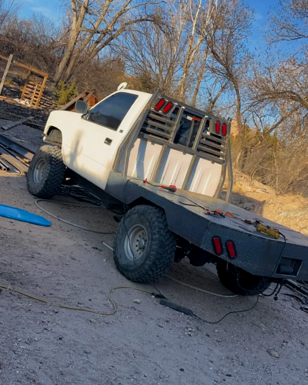 A white pickup truck without a rear cab, sitting on a dirt surface at a construction or work site, with tools and equipment around, and trees with no leaves in the background.