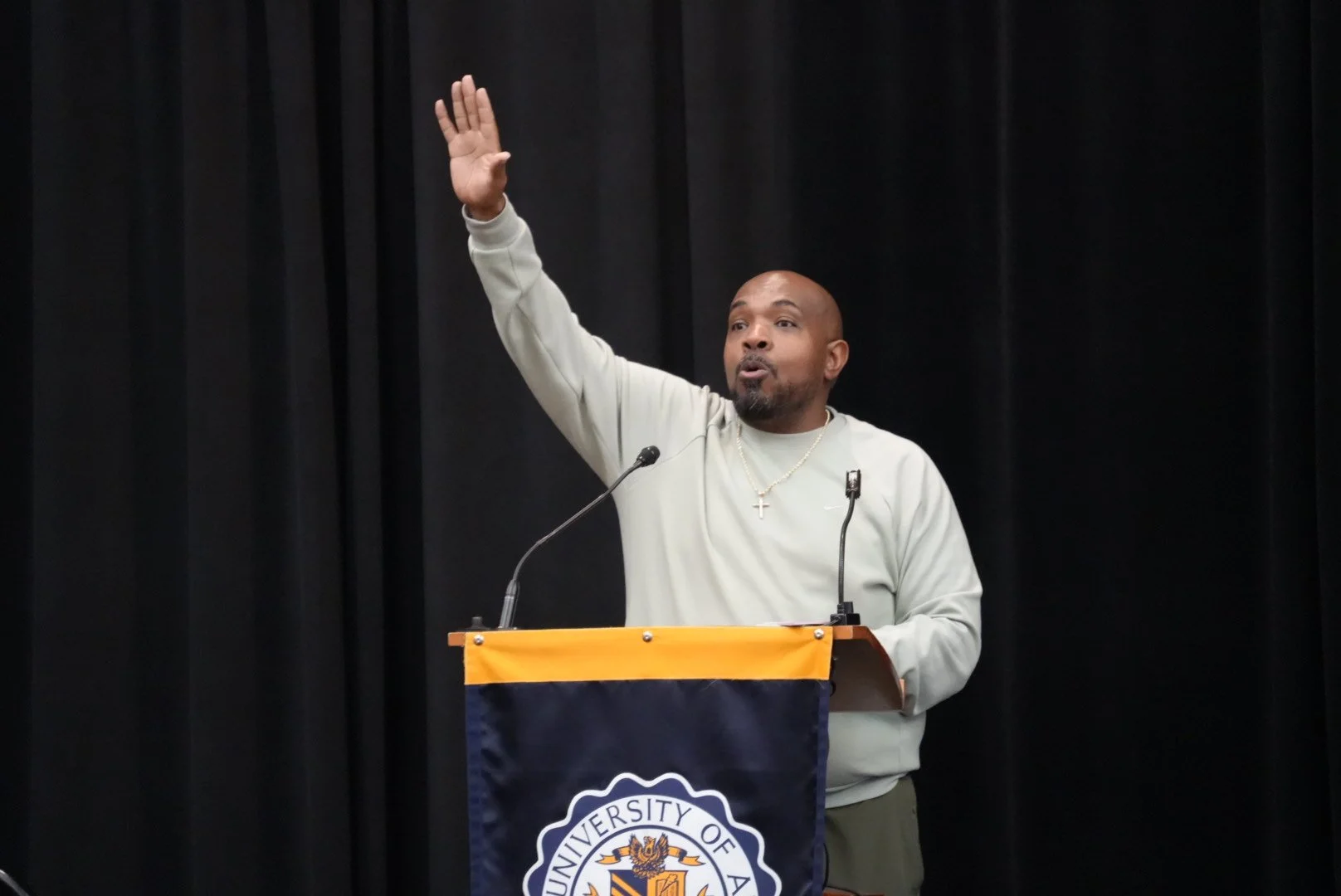 A man speaking at a podium with a University of Alabama banner, raising his right hand, wearing a light-colored sweatshirt, a cross necklace, and a gold chain, against a black curtain background.
