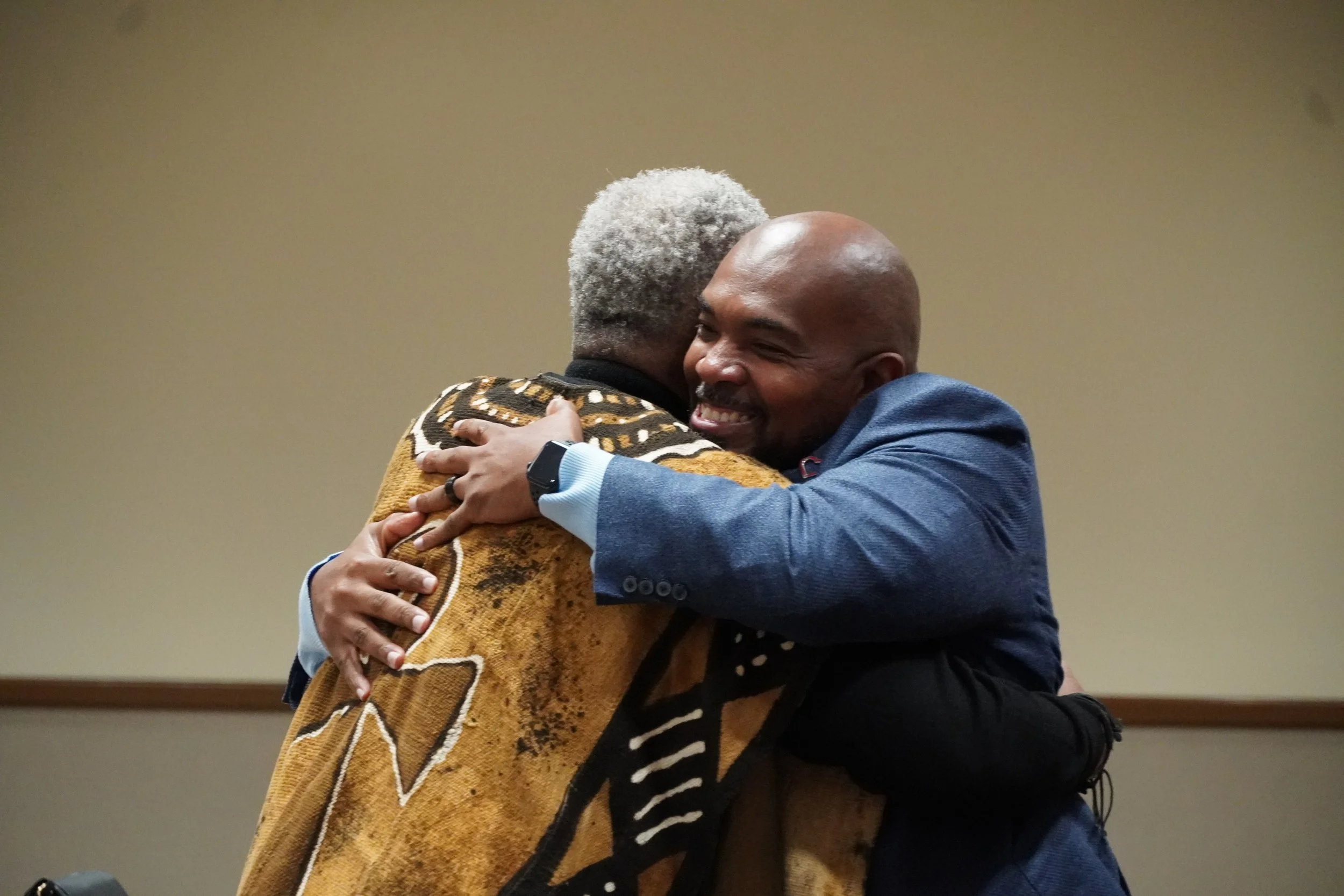 Two men hugging warmly, one facing away with gray hair and patterned sweater, the other smiling with a bald head and navy blazer.
