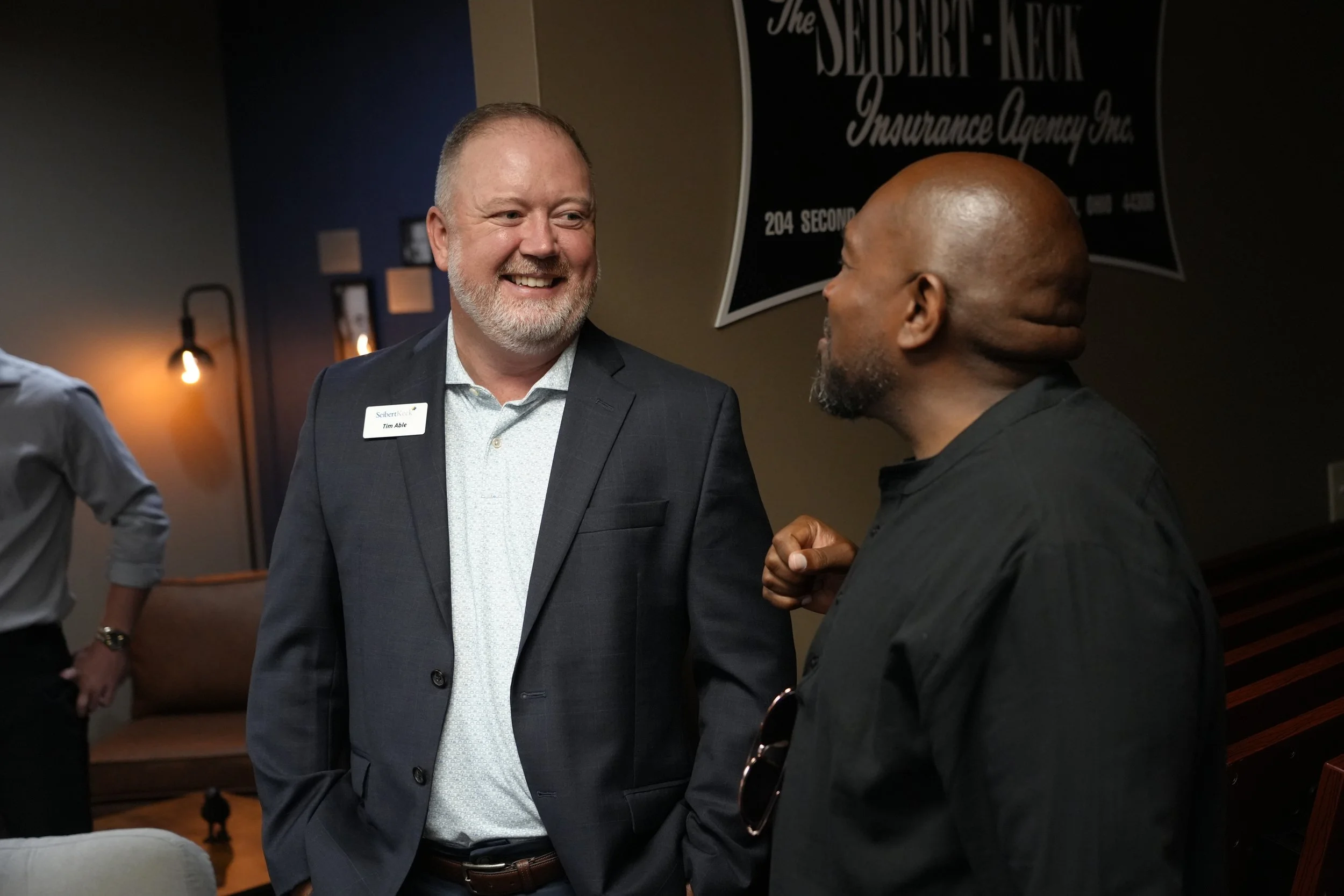 Two men are talking at an event, one wearing a business suit with a name tag that reads 'Tim Able' and the other with a bald head and beard. They are smiling and standing near a sign that reads 'The Seibert-Keck Insurance Agency Inc.' in a dimly lit room with wall-mounted lights.