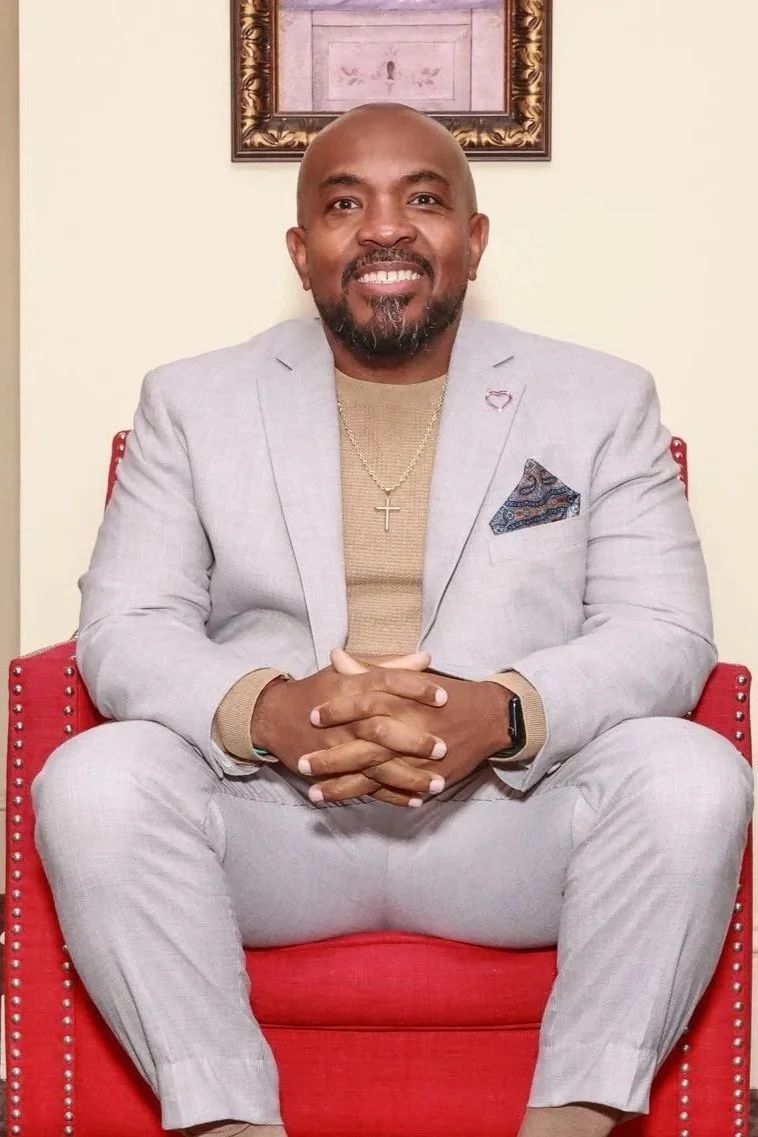 A man sitting on a red chair, smiling, wearing a light gray suit, a beige shirt, a cross necklace, and a pocket square, with a framed picture and cream-colored wall in the background.