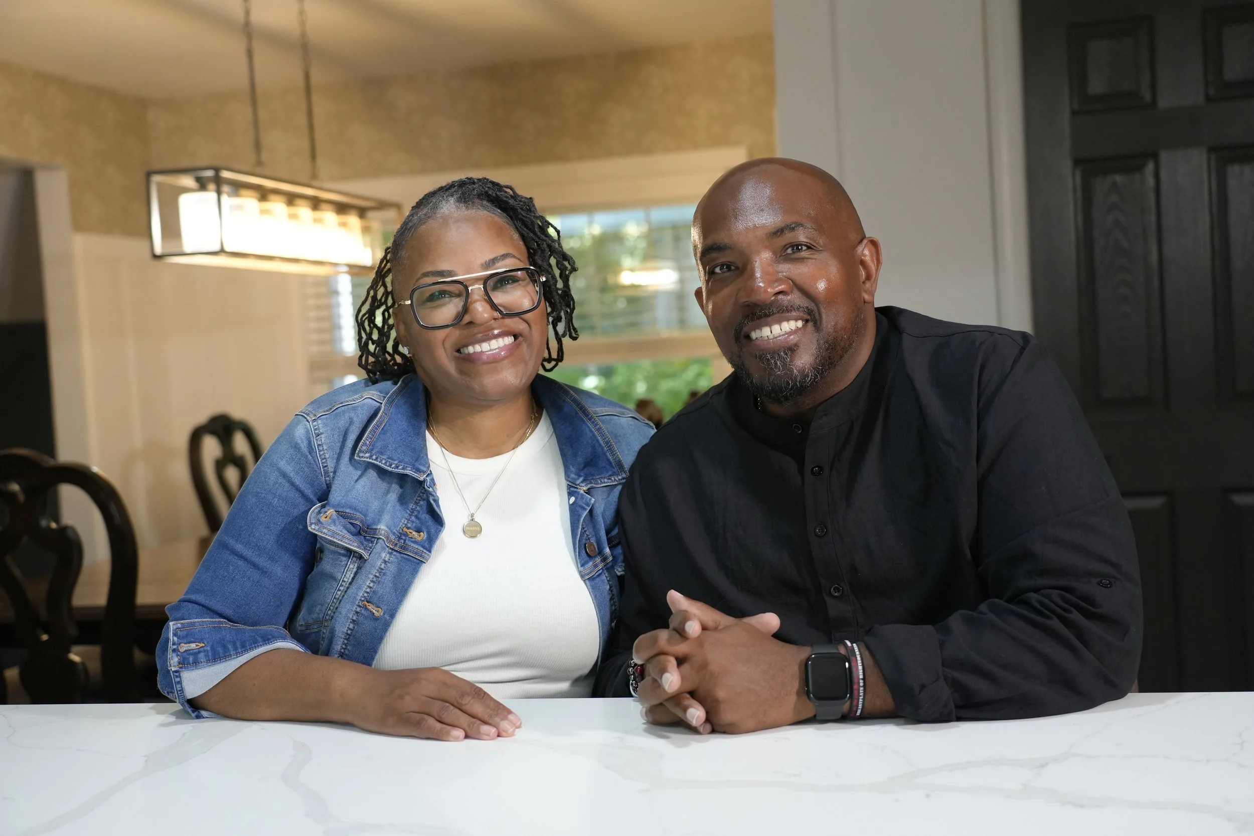 A smiling Black woman with glasses and a Black man with a shaved head and goatee sitting at a white table in a well-lit room.