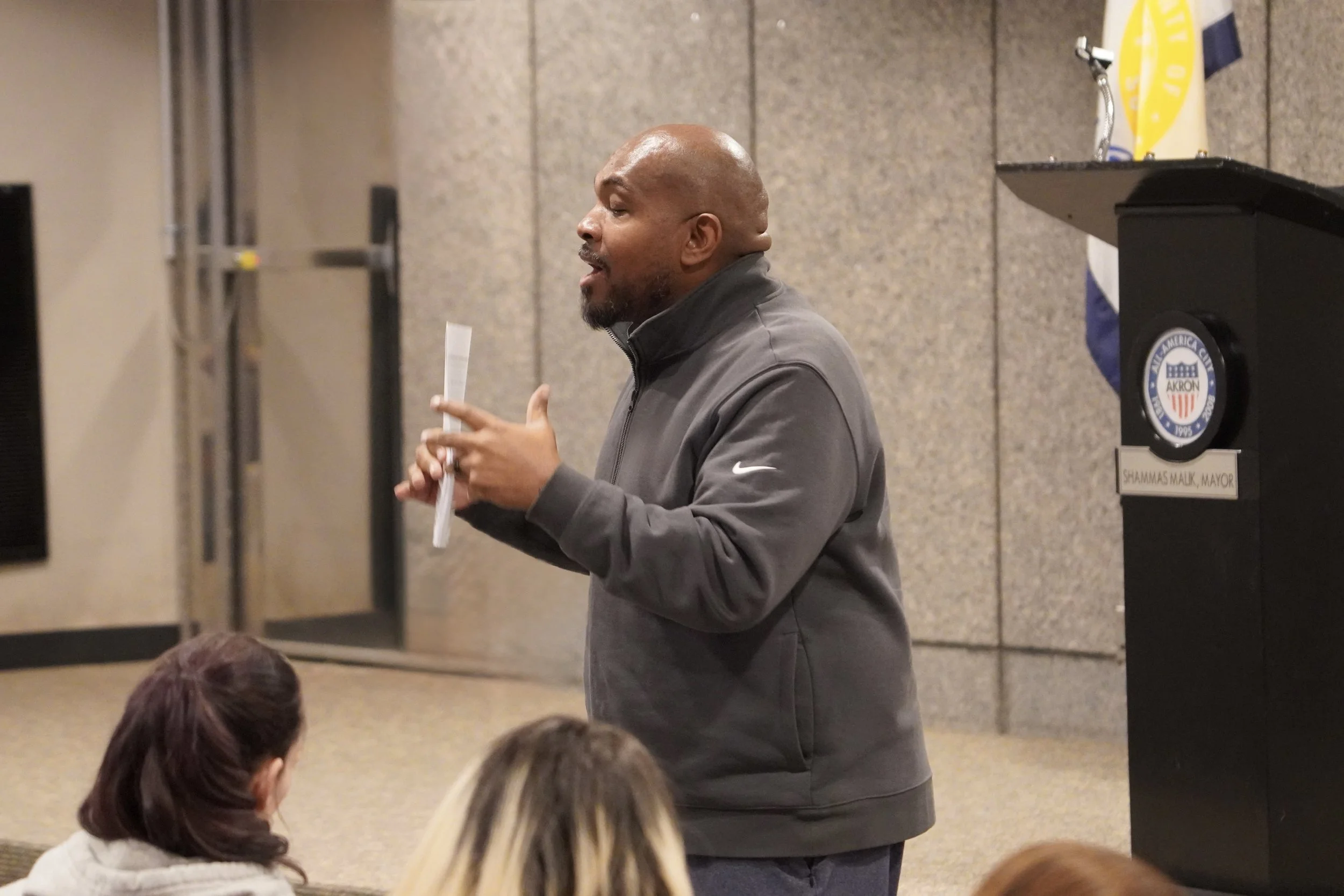 Man speaking to a group of people while holding a paper, at a podium with an Akron, Ohio seal and a yellow flag in the background.