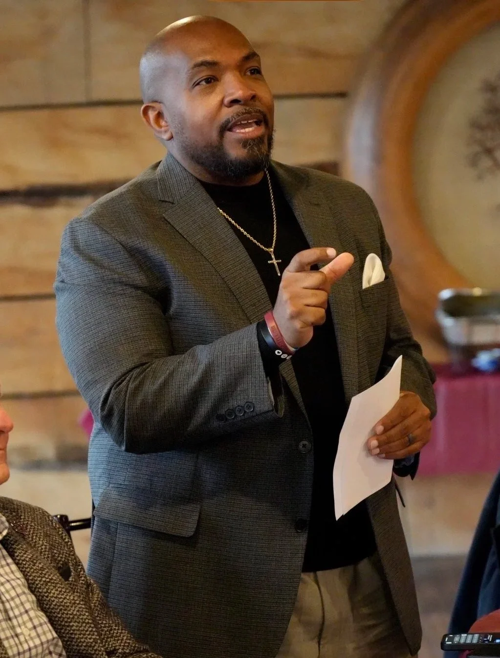 A man wearing a blazer, black shirt, and cross necklace standing and speaking, holding a paper in one hand, with a wooden wall and clock in the background.