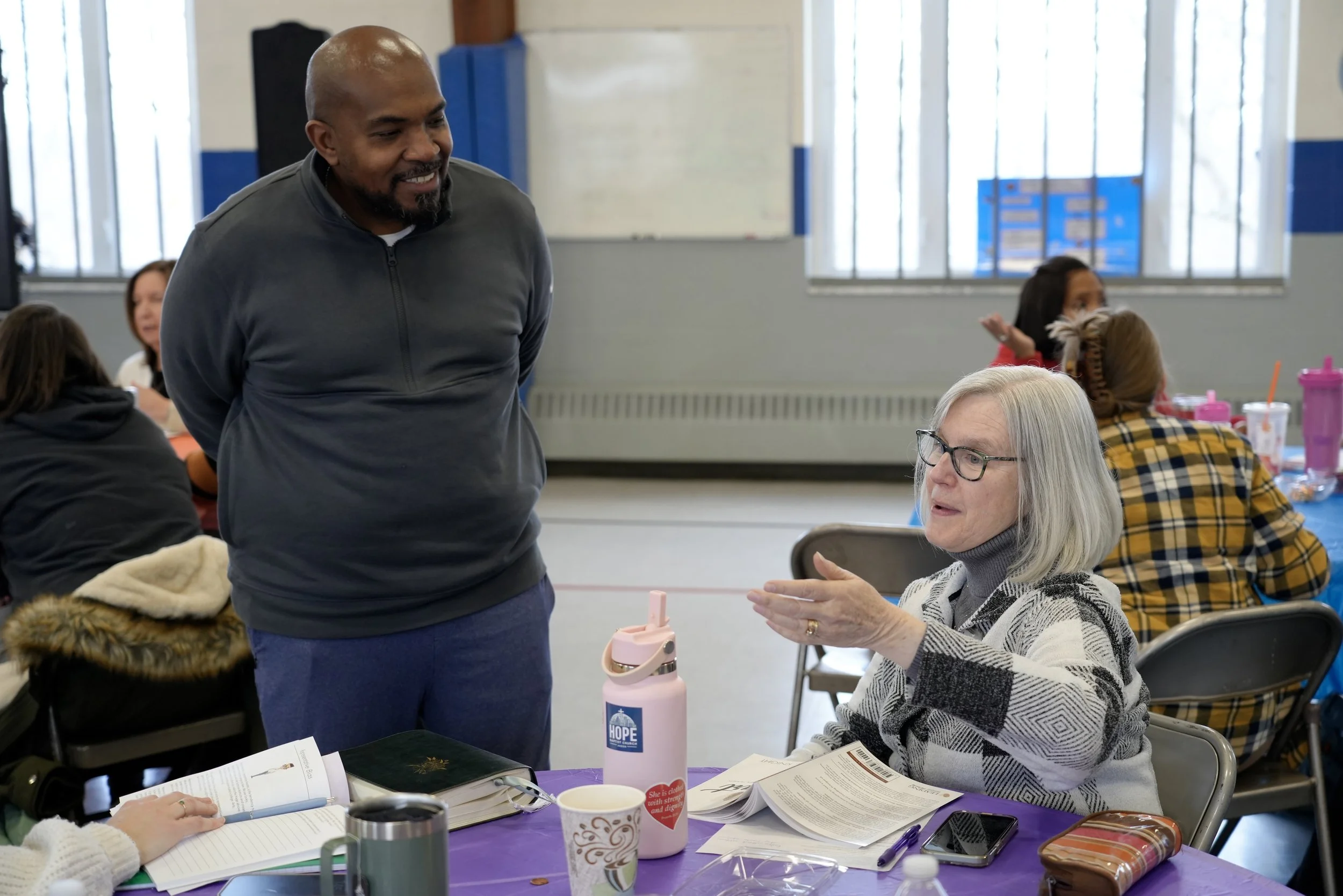 A man is smiling and talking to an elderly woman who is seated at a table in a community center or classroom. The table has papers, a notebook, and a water bottle. Other people are in the background, and large windows provide natural light.