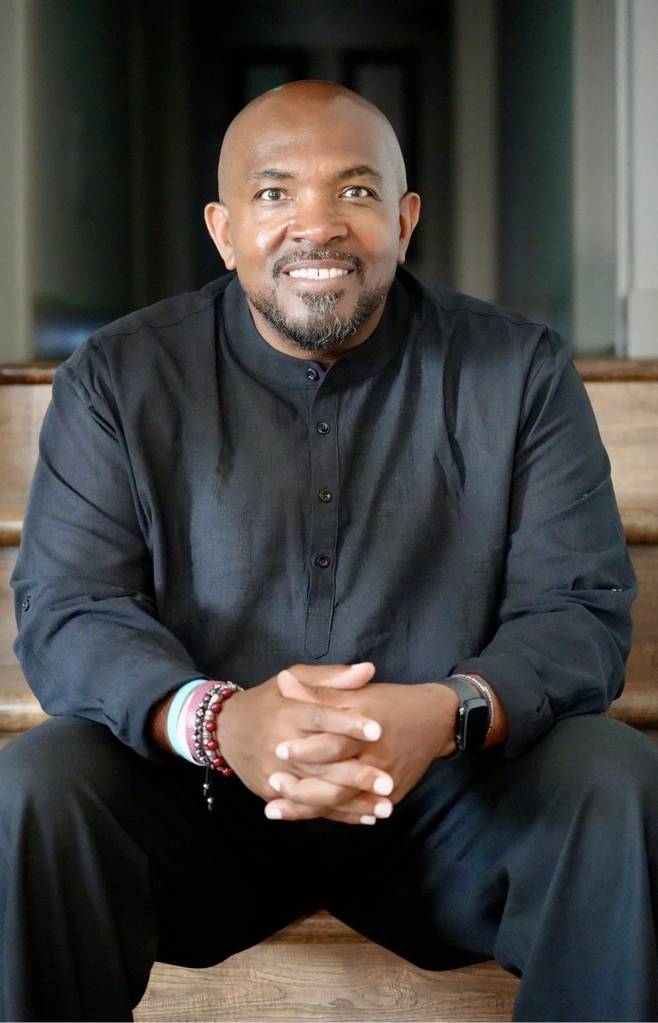 A man sitting indoors at a wooden table, smiling, with his hands folded in front of him. He has a shaved head, a beard, and is wearing a black shirt. He is accessorized with bracelets, a watch, and a fitness tracker.