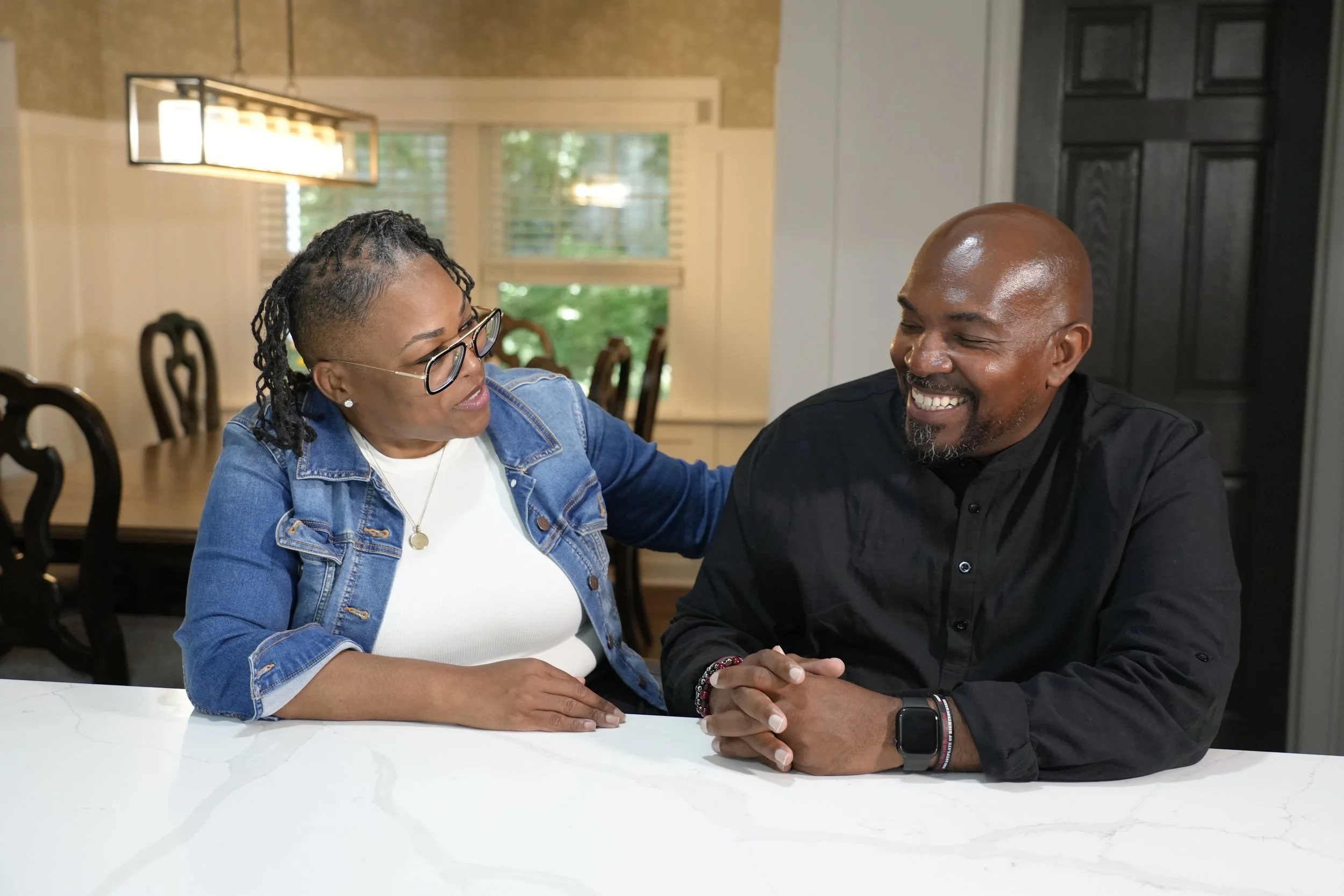 A woman and a man are sitting at a white marble table, engaging in a heartfelt moment. The woman wears glasses, a denim jacket, and jewelry, while the man is bald, smiling, dressed in a black shirt, and wearing a smartwatch. They hold hands and look at each other with affection inside a well-lit domestic setting.
