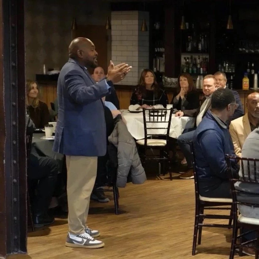 A man in a blue blazer and beige pants speaking to an audience in a restaurant or banquet hall, with several people sitting at tables listening attentively.