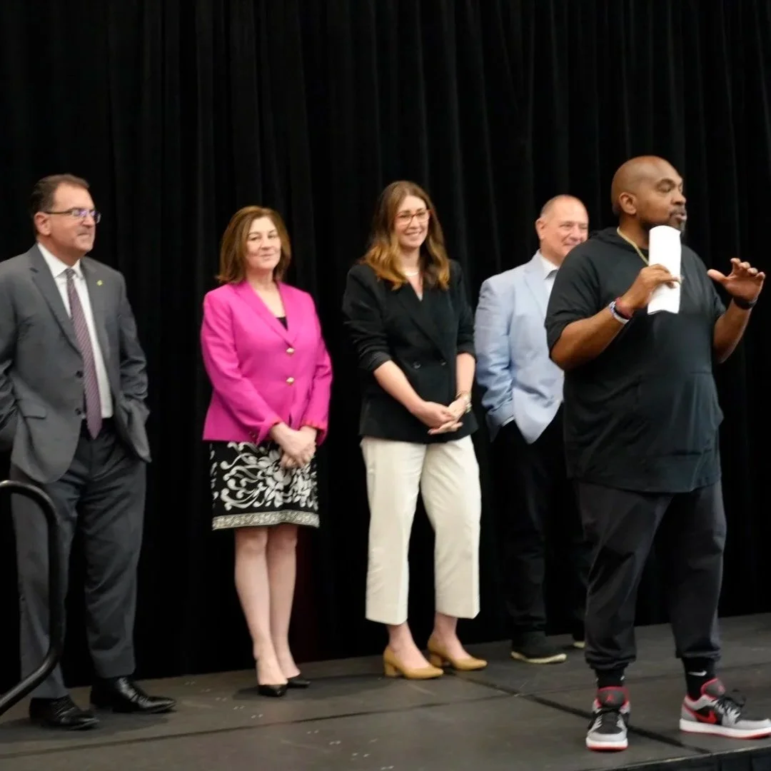 Group of five adults standing on a stage, with a man speaking into a microphone in front of a black curtain.