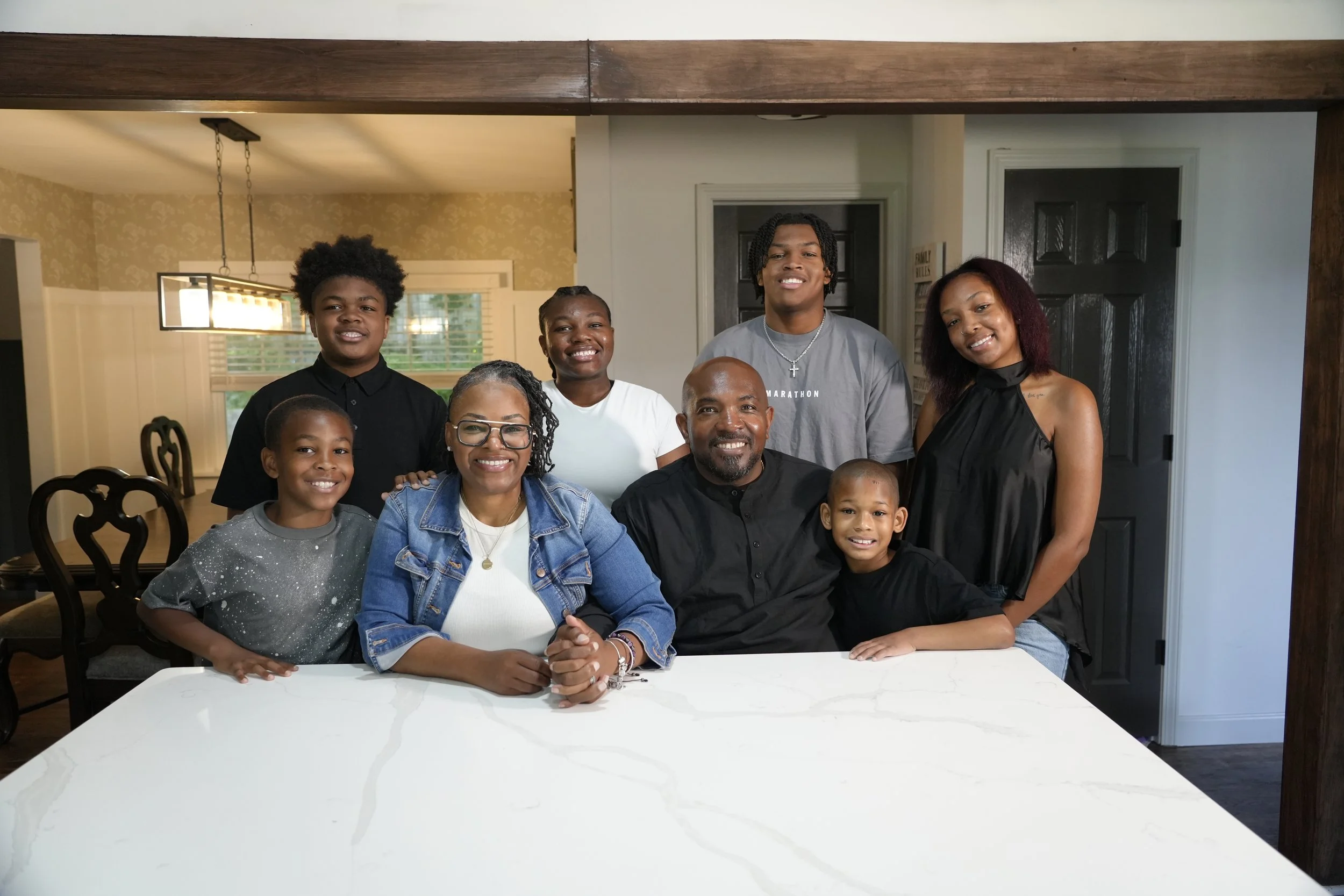 A multi-generational African American family of nine people gathered around a dining table, smiling for a group photo inside a home with a modern kitchen and dining area.