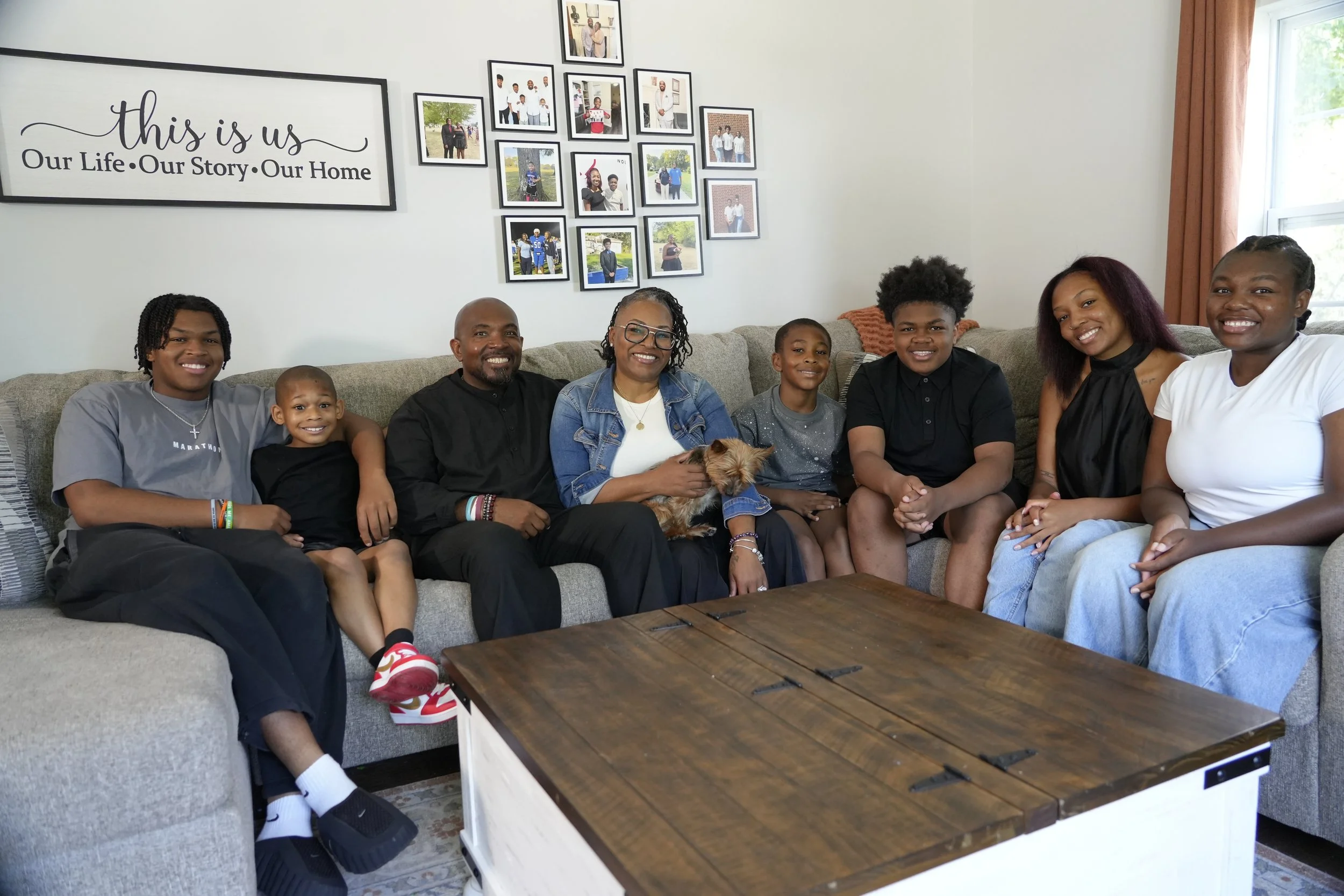 A multi-generational family of nine people and a small dog sitting on a gray couch in a living room, smiling for a photo. There is a wall with framed photos and a large sign that reads 'this is us Our Life Our Story Our Home.' A window with orange curtains is visible on the right.