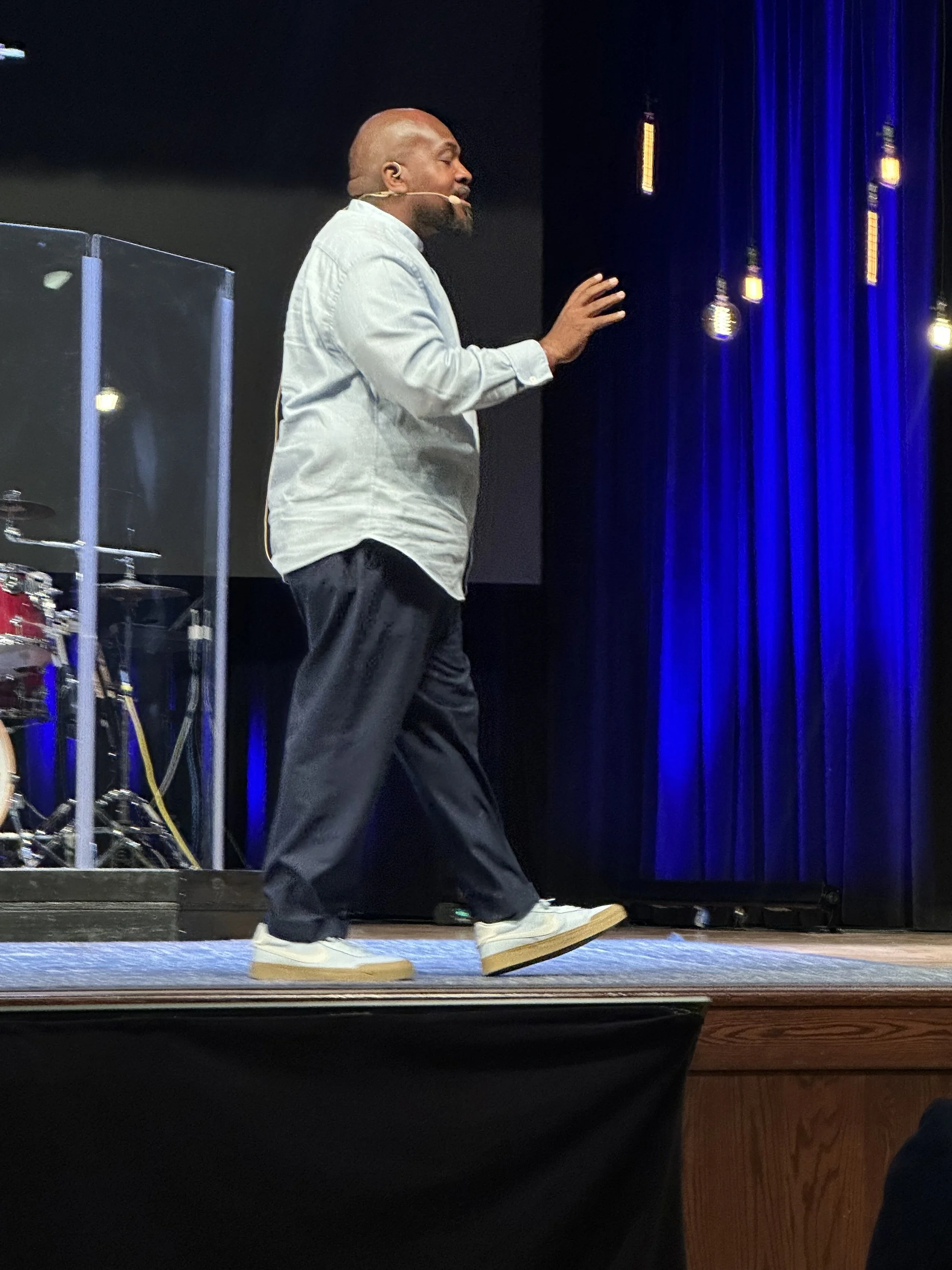 A man with a bald head and beard performing a slide on a skateboard on a stage with blue curtain backdrop and hanging lights, wearing a light-colored shirt, dark pants, and white sneakers.