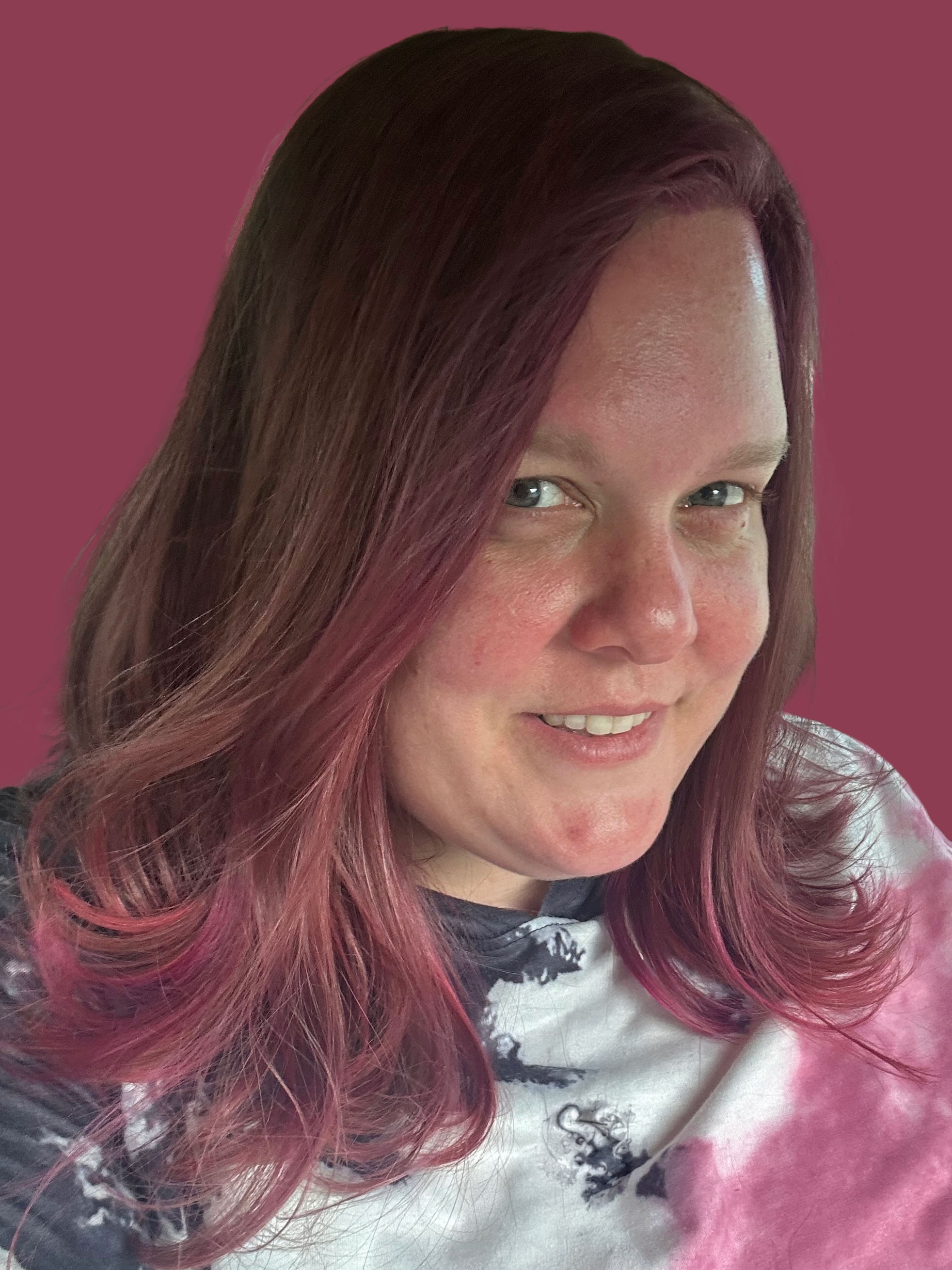 A woman with long pinkish-purple hair smiling at the camera, wearing a patterned top, with a solid pink background.