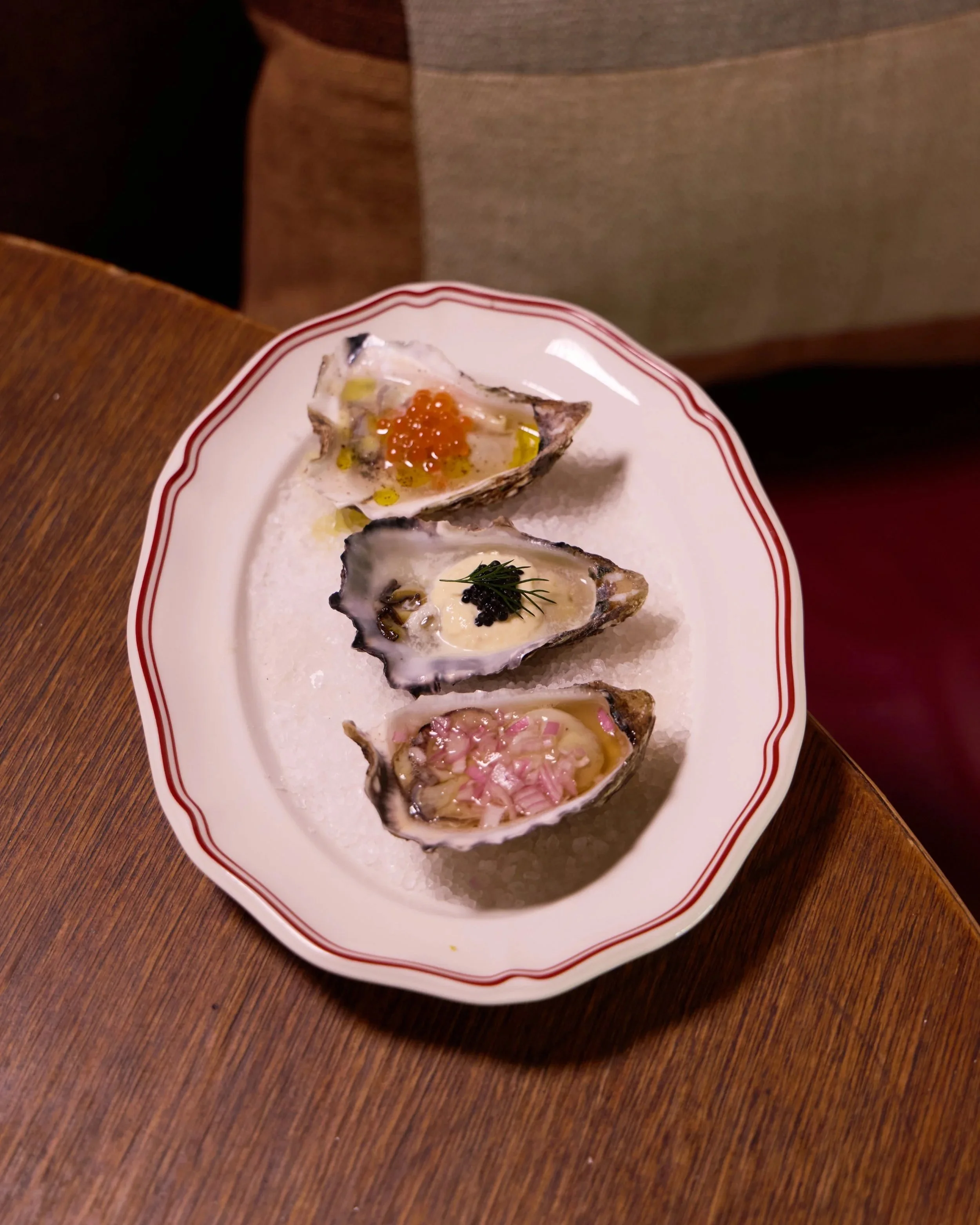 Three half-shell oysters on a white plate with red decorative rim, topped with toppings, on a wooden table.