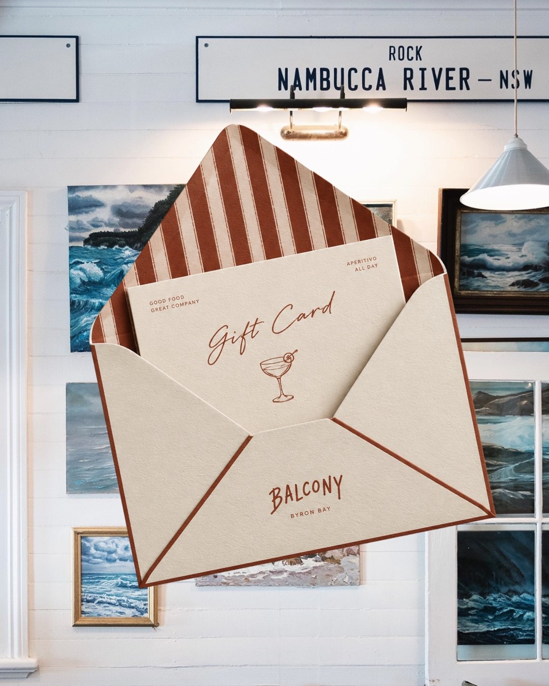 A gift card in an envelope labeled 'Balcony Byron Bay', inside a room with ocean-themed artwork and a sign indicating Nambucca River, NSW.