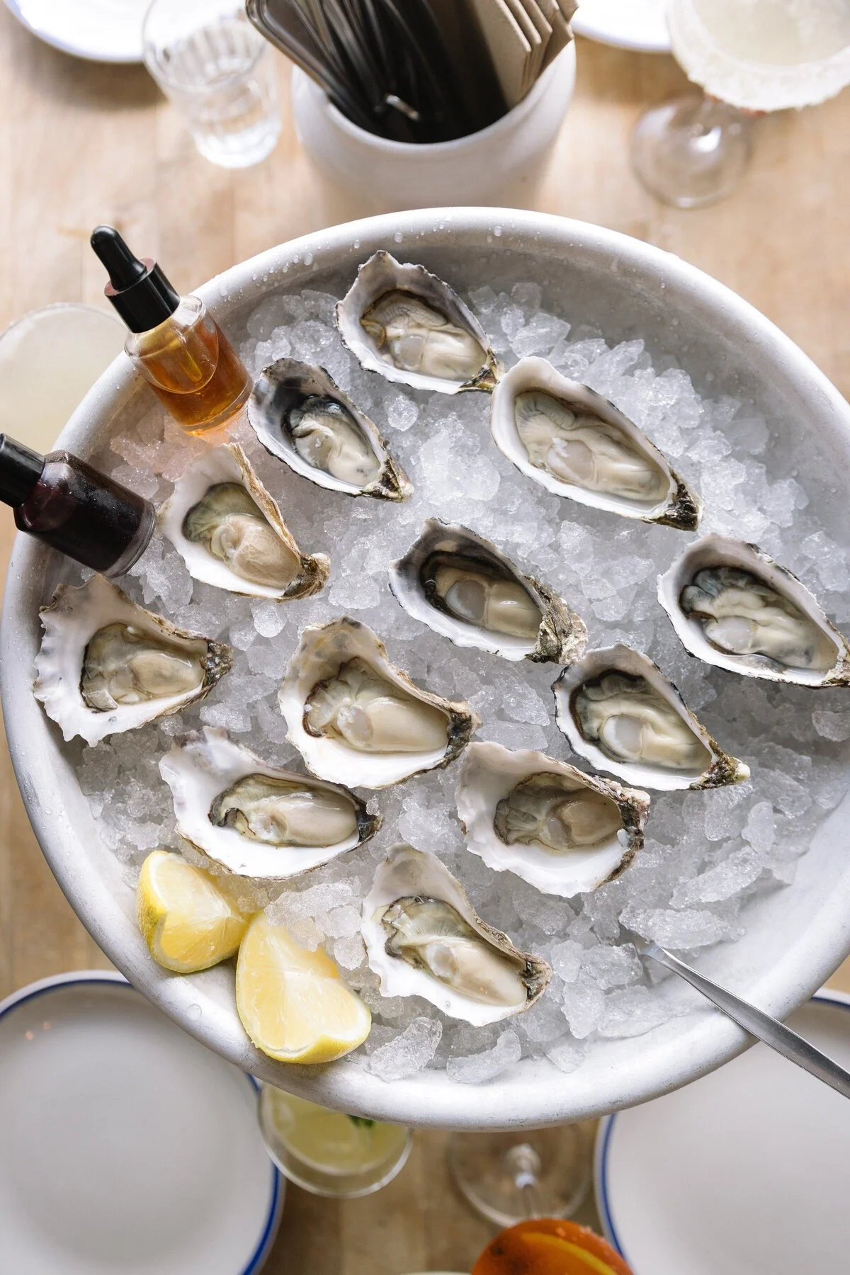 A silver ice bucket filled with crushed ice, fresh oysters on the half shell, lemon wedges, and small bottles of hot sauce and cocktail sauce. Glasses of beverages and utensils are visible in the background.