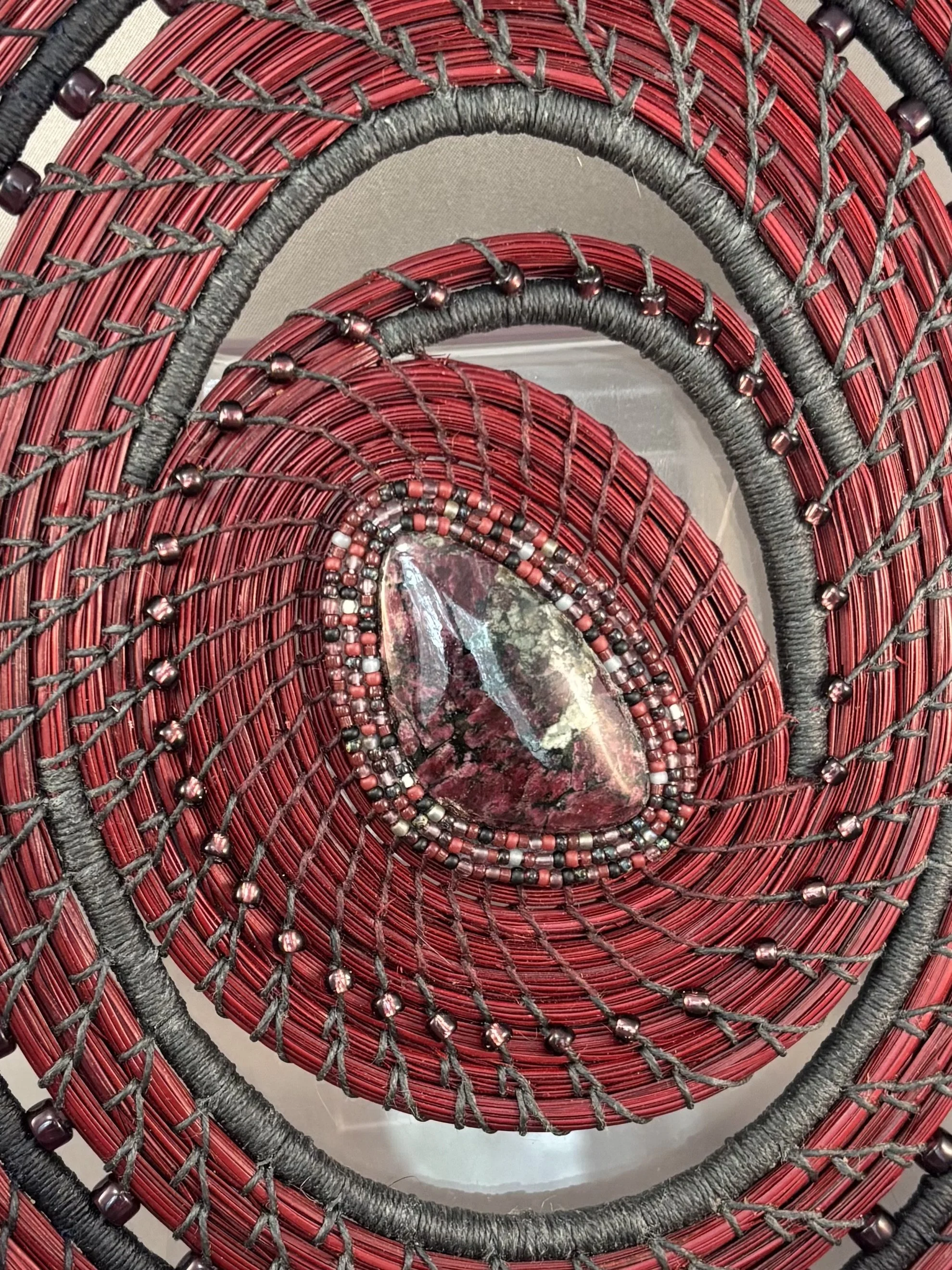 Detail of eudialite cabochon surrounded by three rows of colourful seed beads, coiled red-dyed pine needles