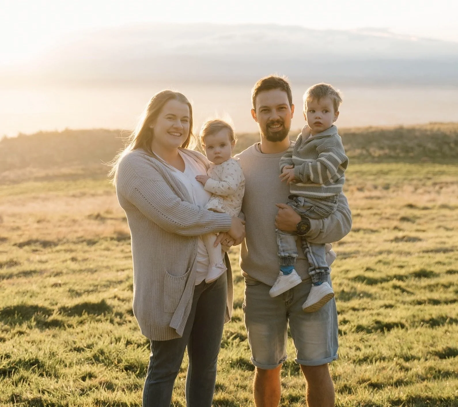A family of four standing outdoors during sunset, smiling and holding two young children, on a grassy field with hills in the background.