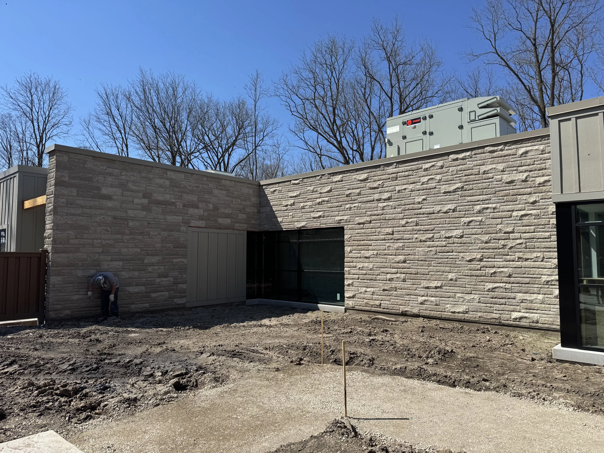 Construction site with a dirt ground, a brick wall and house exterior with large glass windows, and HVAC units on the roof, under a clear blue sky with leafless trees in the background.