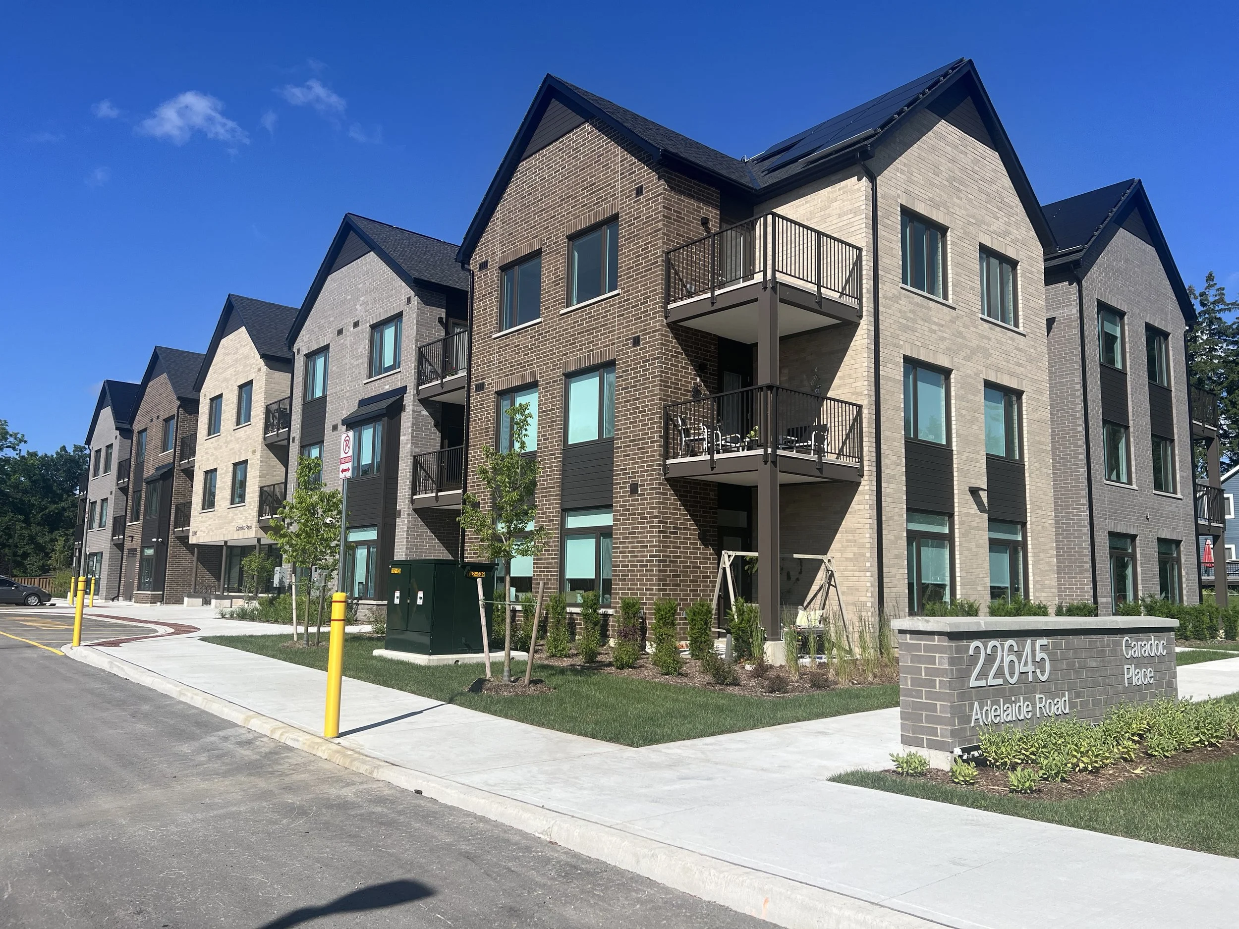 Modern multi-story residential building with brick and siding exterior, balconies, and large windows, situated on a street with sidewalk, small landscaped trees and bushes, and street signs showing the address 22645 Adelaide Road.