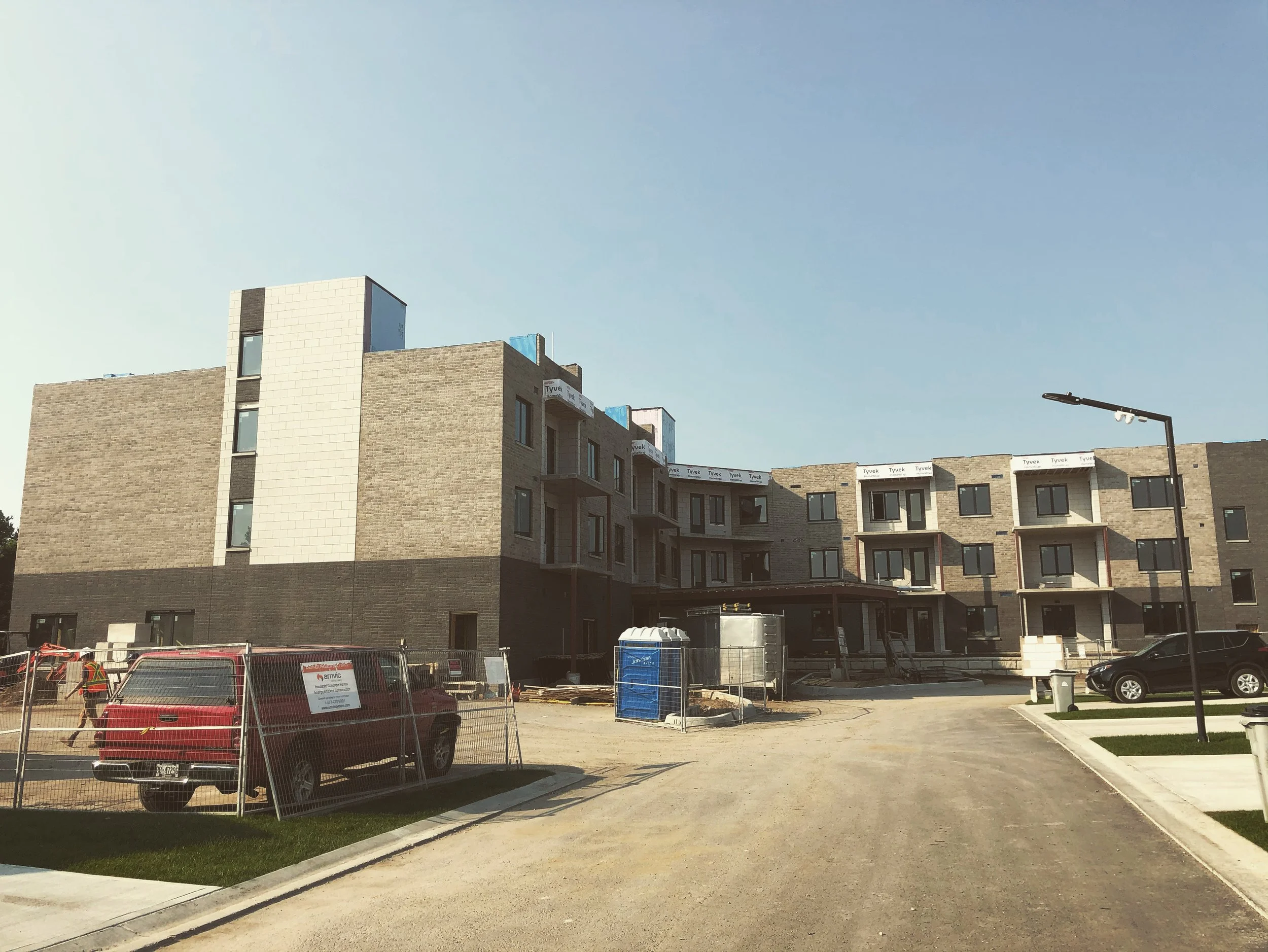 A construction site with partially built residential buildings, parked cars, construction barriers, and a streetlamp under a clear blue sky.