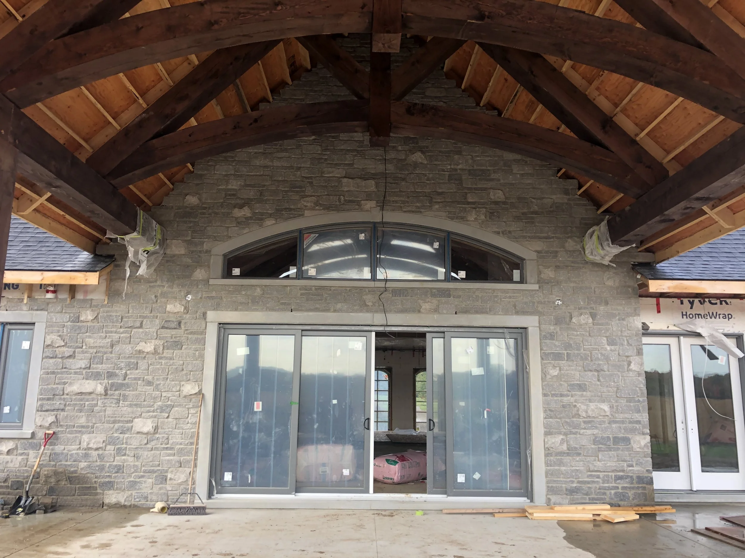 The image shows an unfinished building or house exterior with a stone brick wall, large sliding glass door, and a vaulted wooden ceiling with exposed beams under construction.
