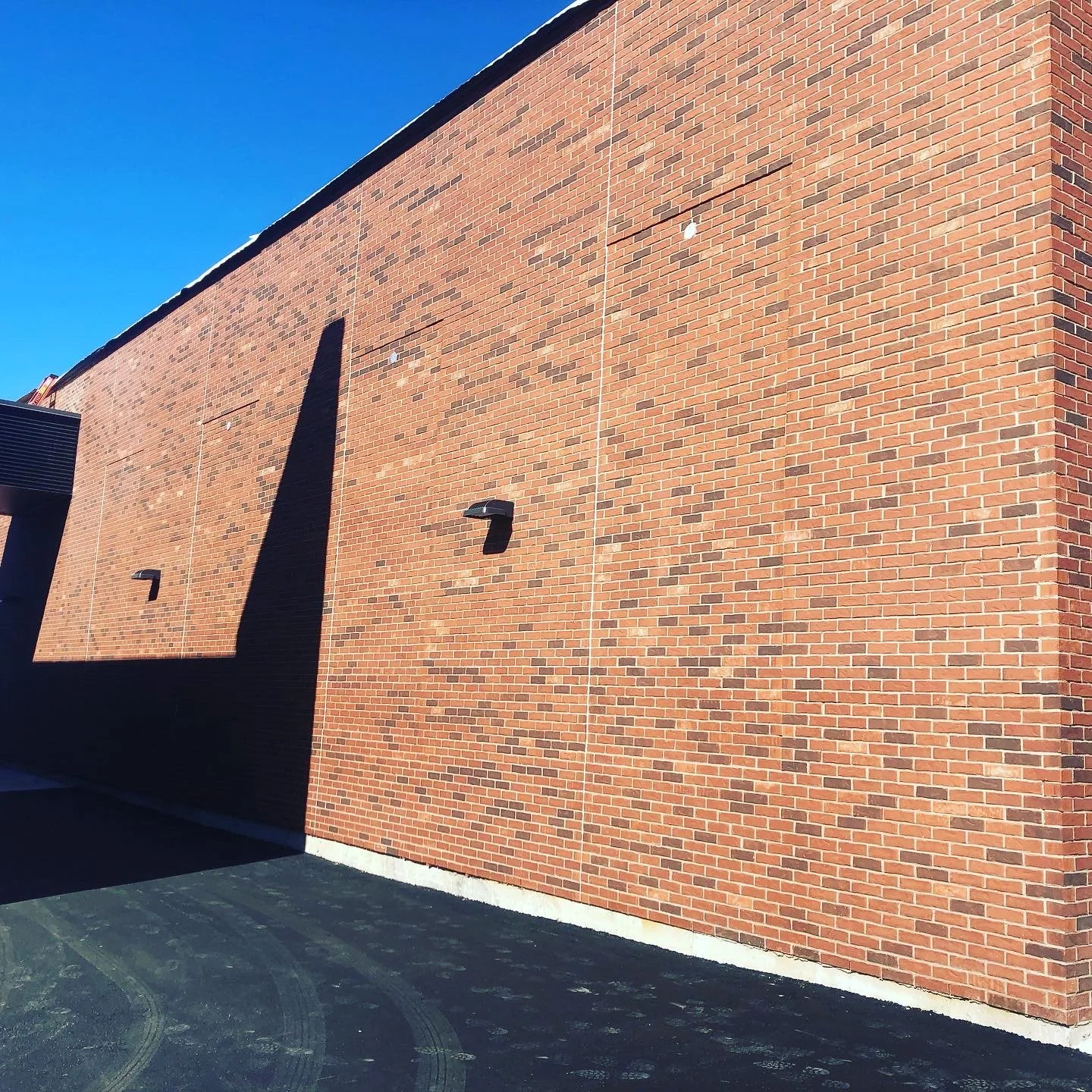 A large brick wall on a building with top edge visible, with two outdoor lights mounted on it, and shadows cast on the wall from nearby objects, under a clear blue sky.