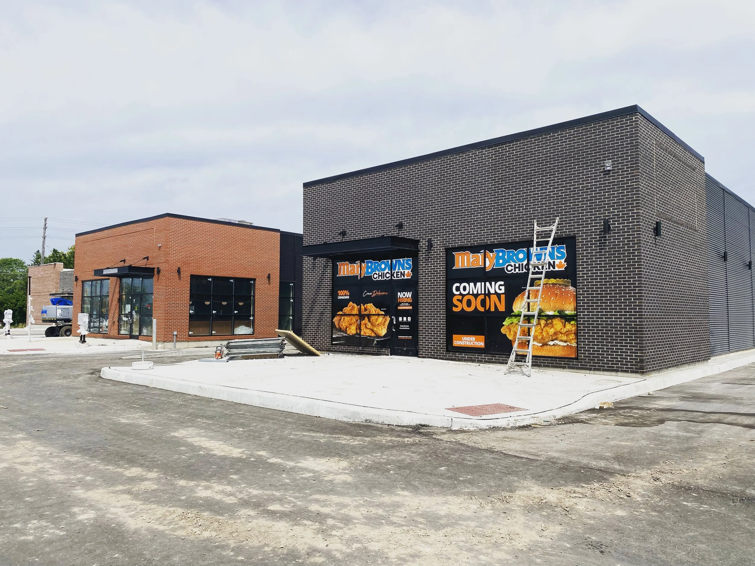 Construction site with a new Mary Browns Chicken restaurant under construction, featuring a black and red brick facade, signs indicating the upcoming opening, and construction equipment, with an overcast sky in the background.